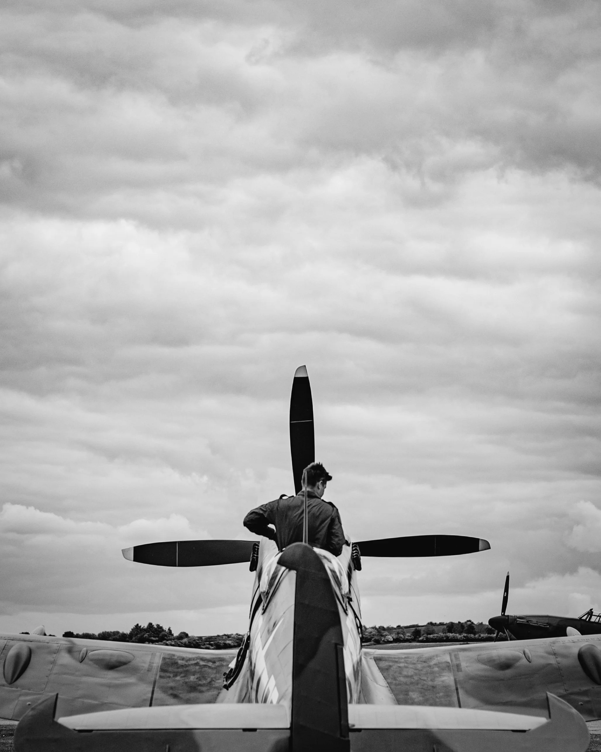 A person is sitting on the tail of a small aircraft, facing away, with the aircraft's tail fin and wings visible against an overcast sky.