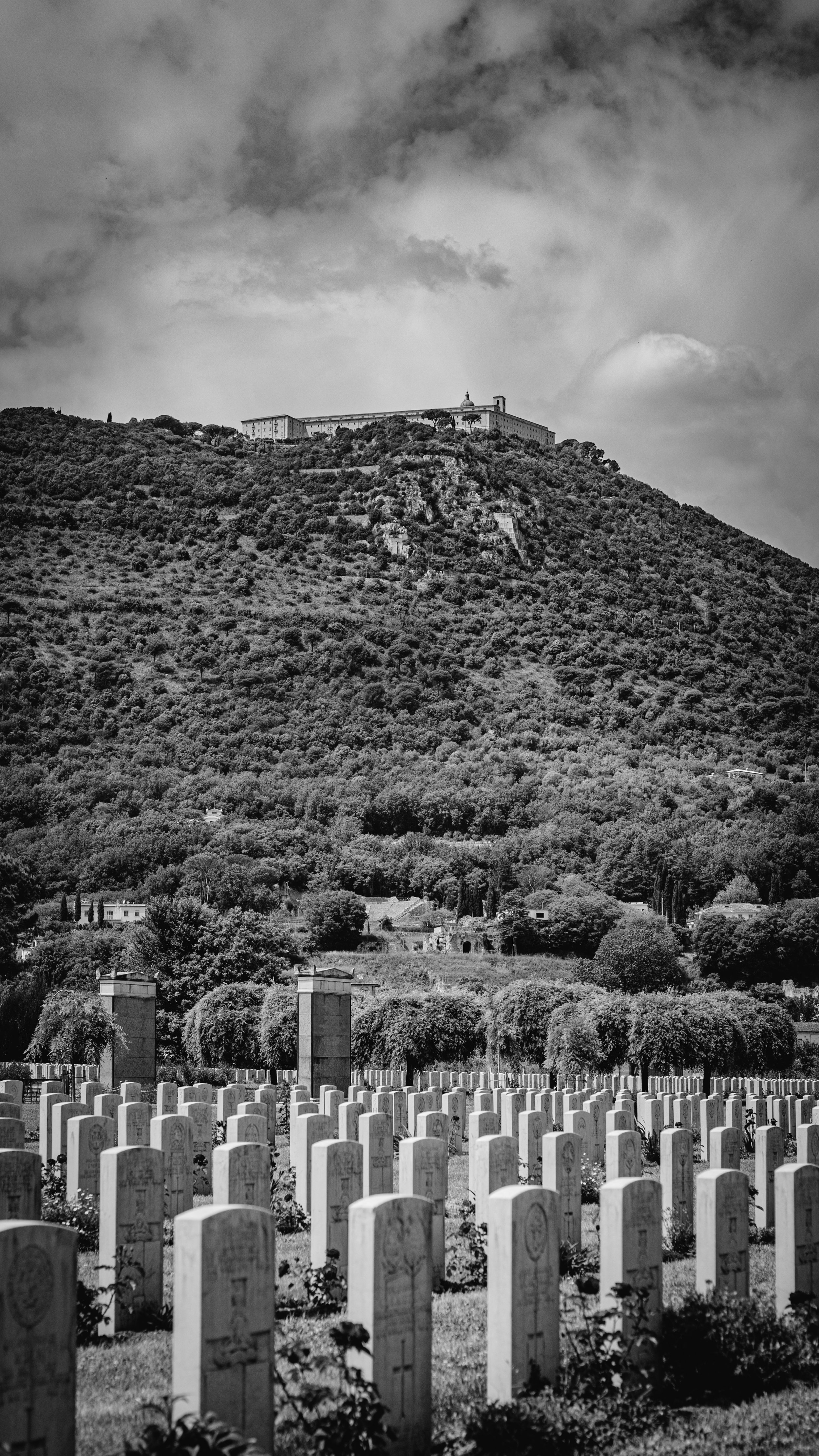 Black and white photo of a military cemetery with rows of gravestones in the foreground, a lush hillside with trees and buildings on the hill, and a cloudy sky above.