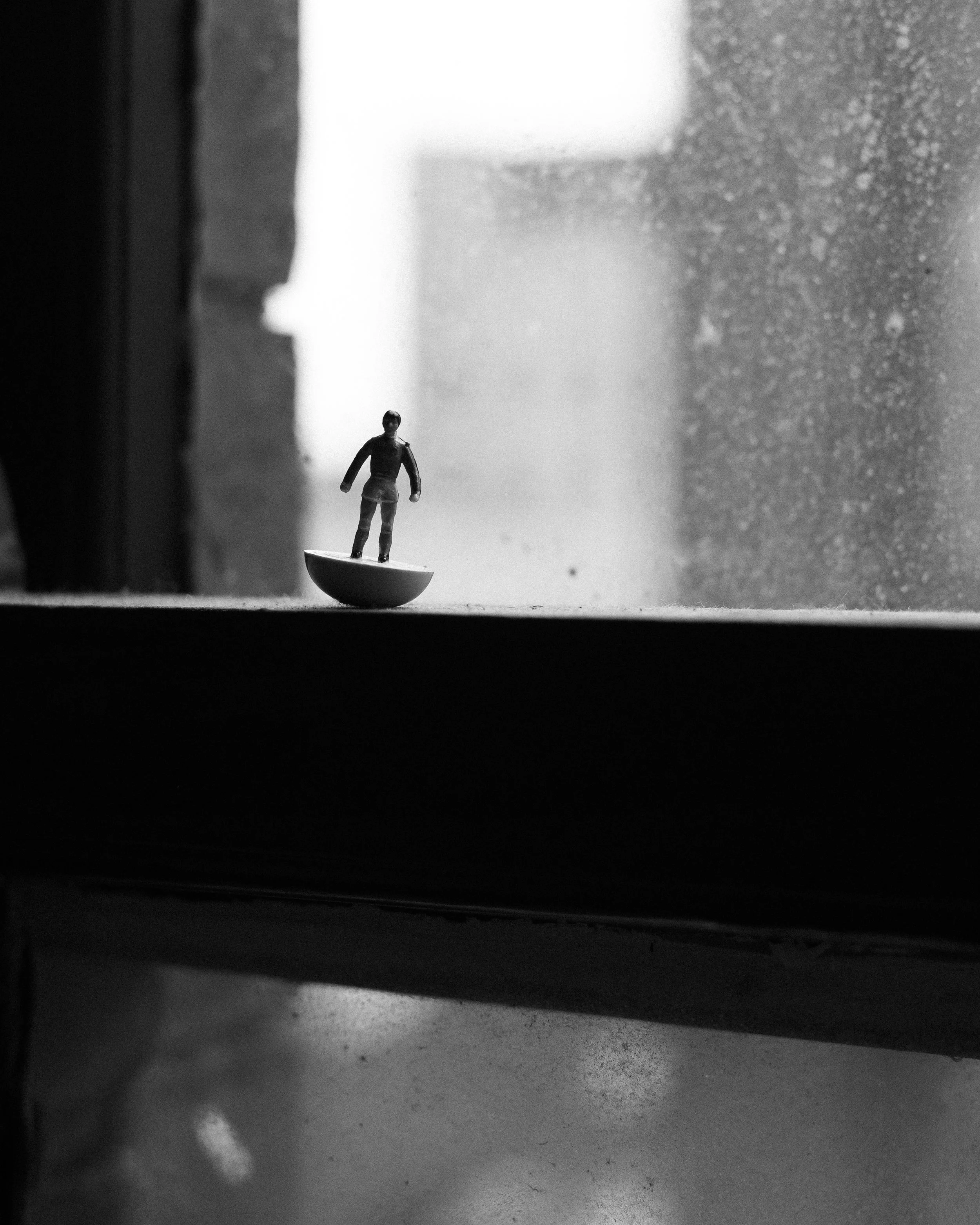 A small toy figure of a woman standing on a platform, with a window and a textured wall in the background.