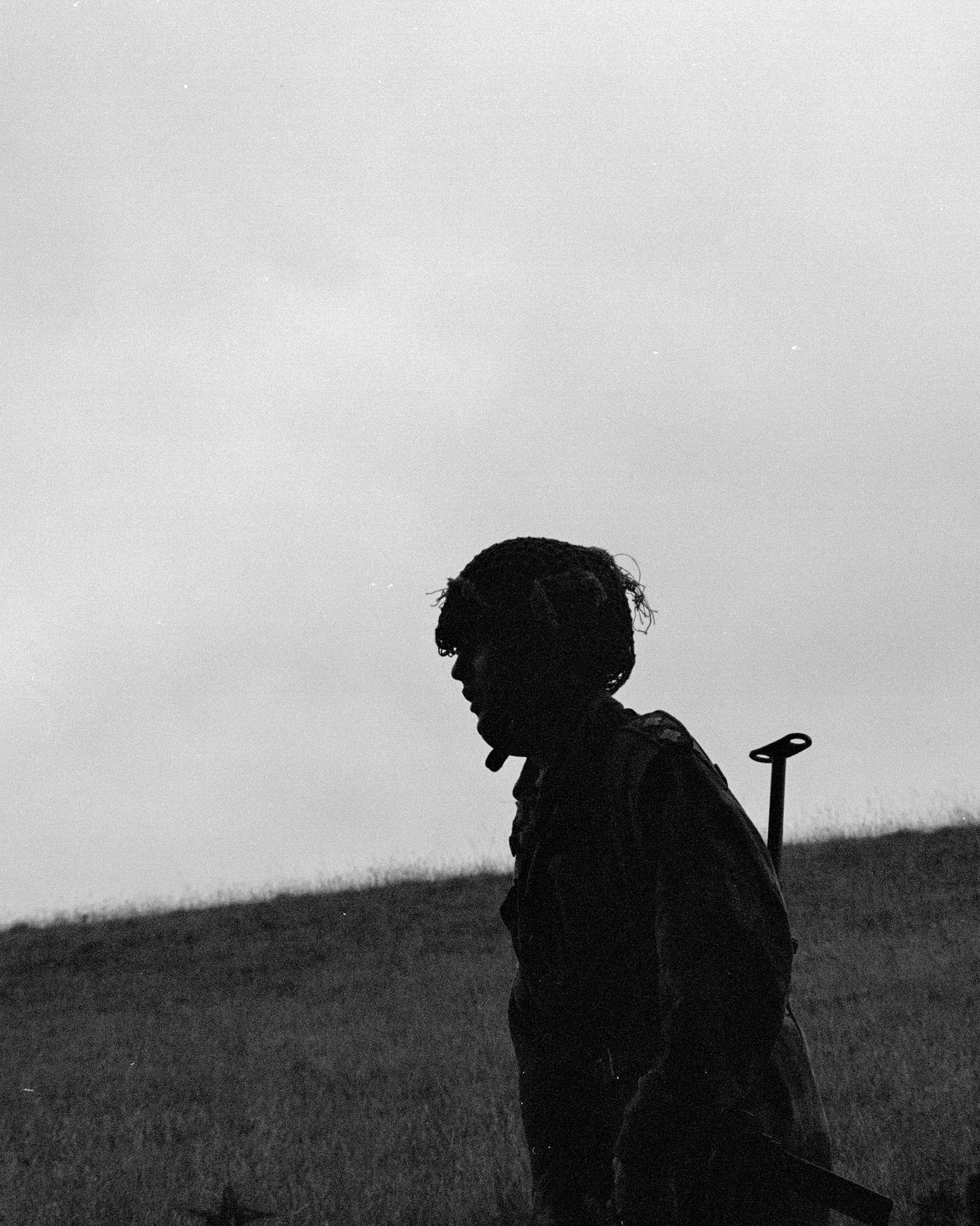 A silhouette of a person with curly hair walking outdoors at dusk, carrying a backpack and a walking stick, with an open landscape and a cloudy sky in the background.