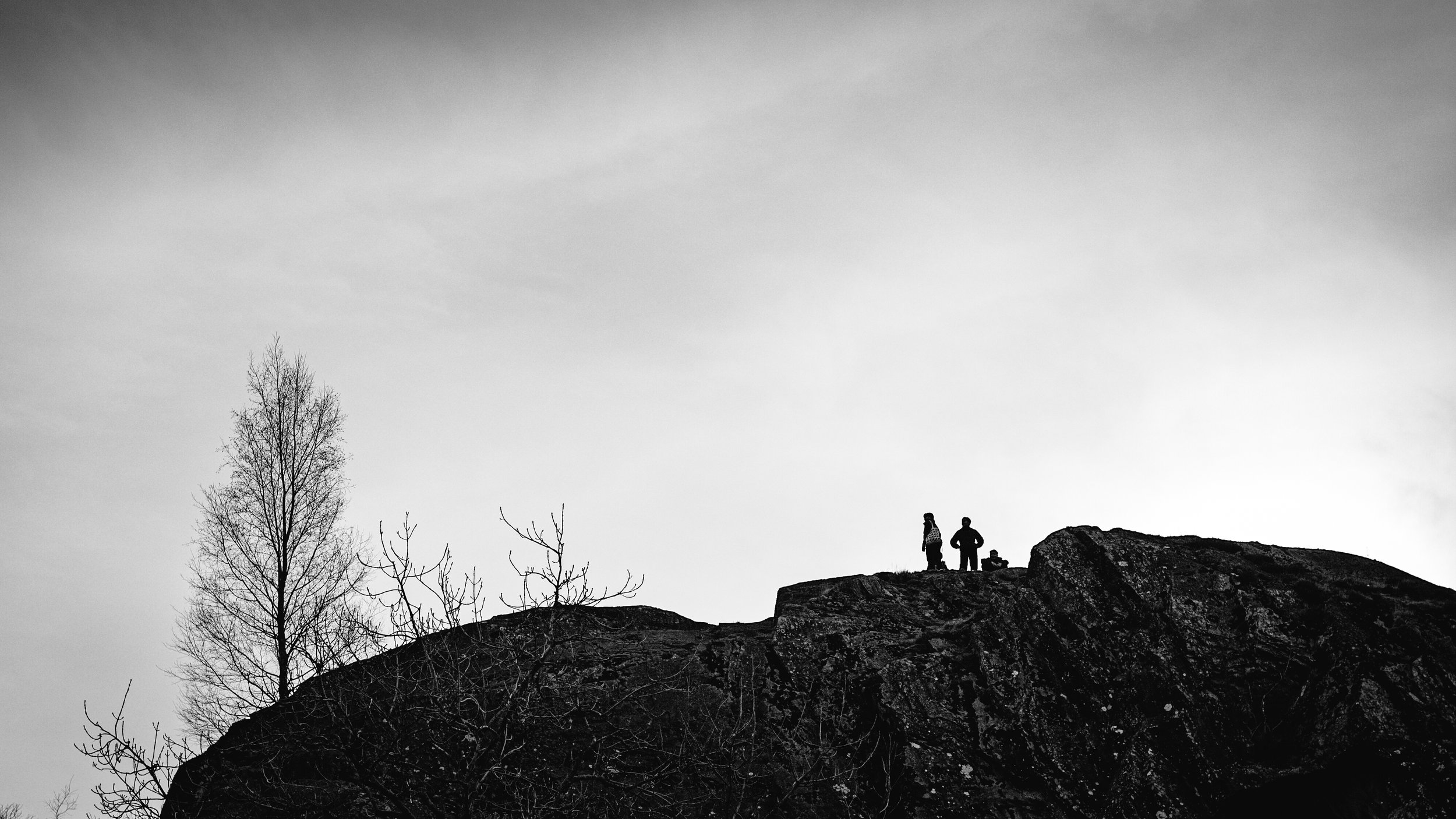 Silhouette of two people standing on a rocky hilltop, with leafless trees and a cloudy sky in the background.