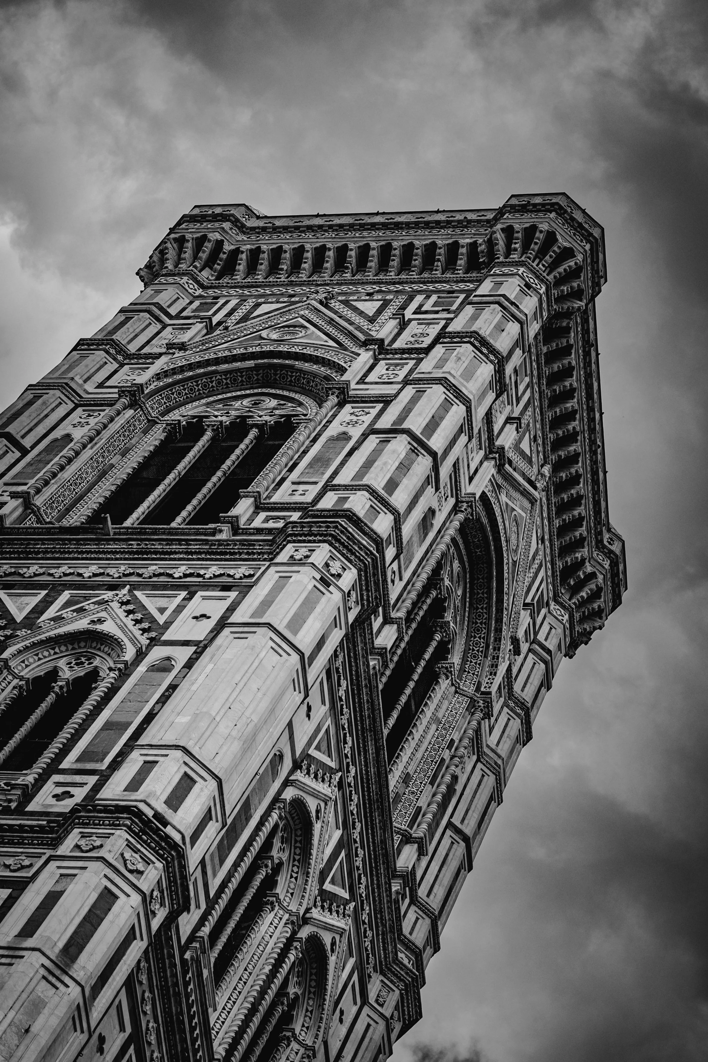 Black and white photograph of a tall, ornate Gothic-style tower with detailed architectural features, set against a cloudy sky.