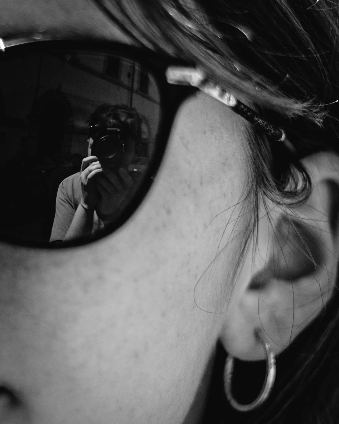 A close-up black-and-white photo of a person's face wearing sunglasses, with the person's reflection visible in the sunglasses. The reflection shows the person taking a photo with a camera, and their hand gripping the camera. The person has dark hair