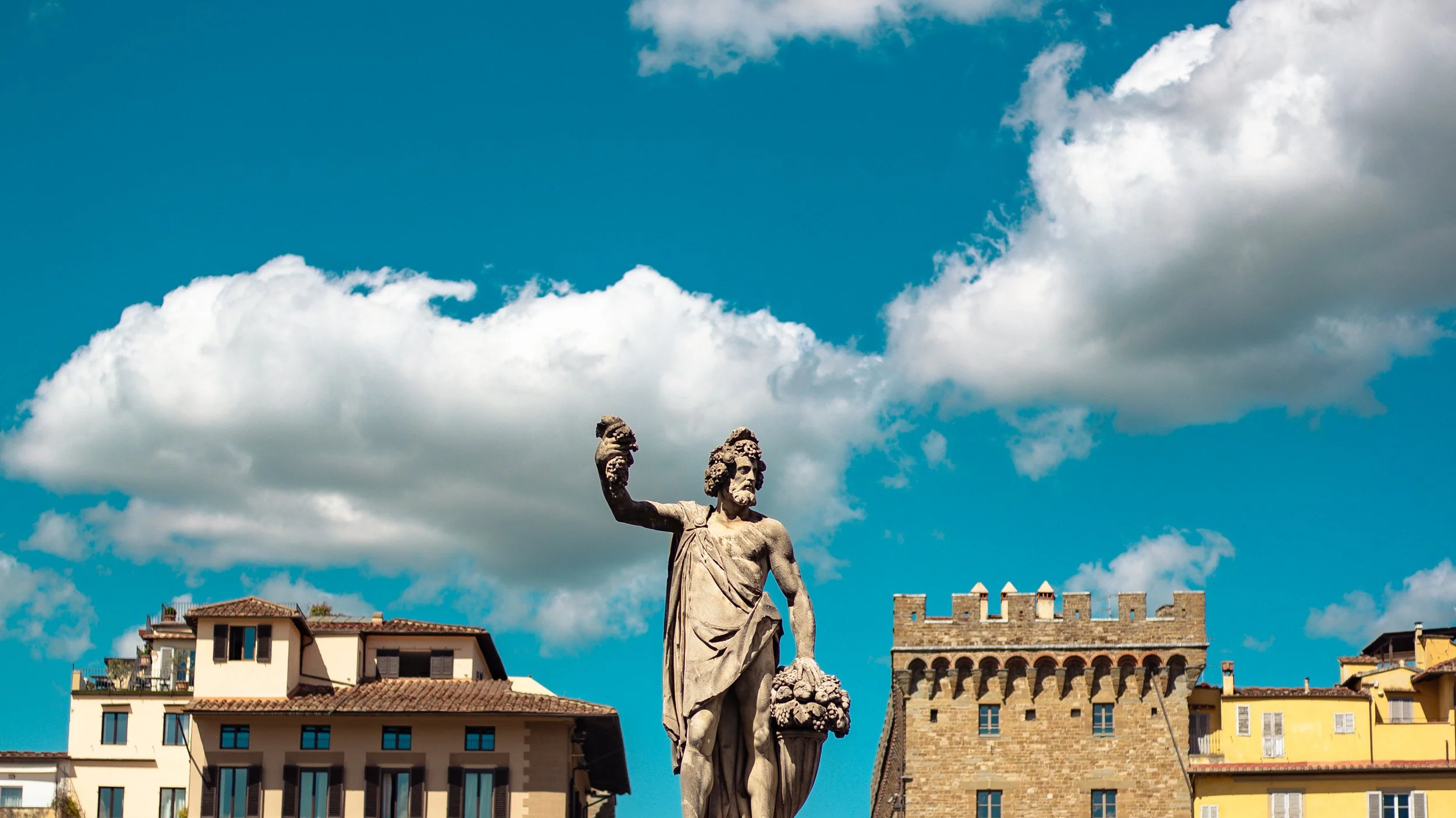 A stone statue of a bearded man holding a bundle of grapes and a staff, set against a background of modern buildings, a stone tower, and a partly cloudy blue sky.