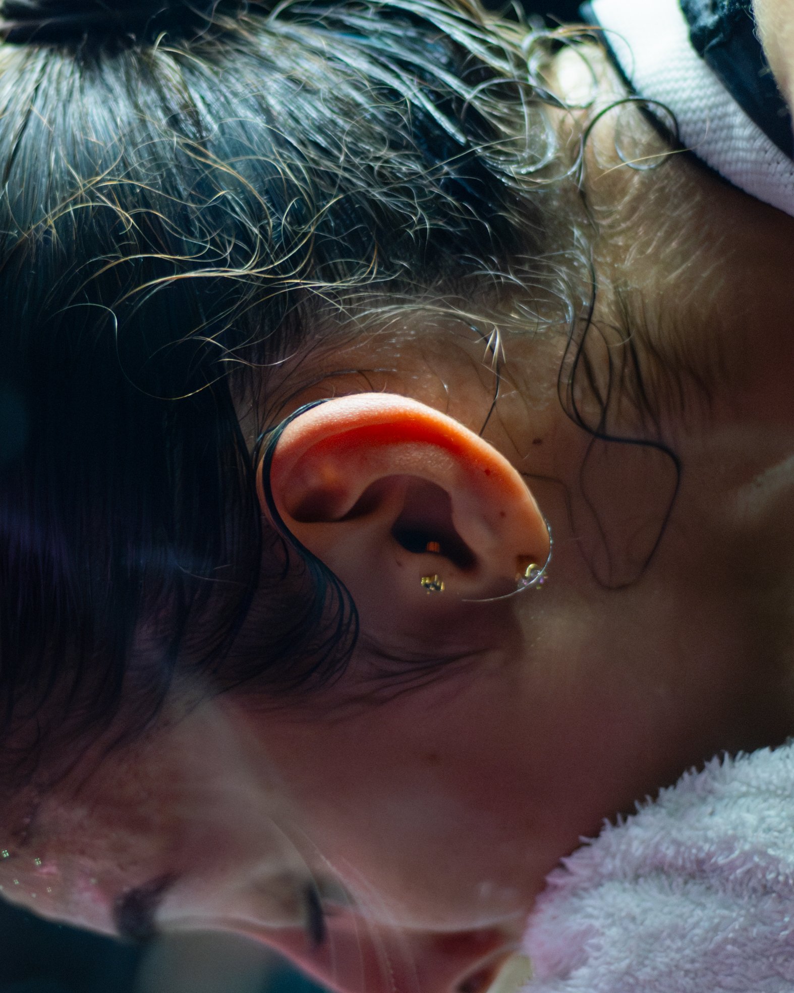Close-up of a person's head with damp black hair, an ear with multiple earrings, and skin with some acne. The person appears to have just washed their hair and is lying down with a towel nearby.