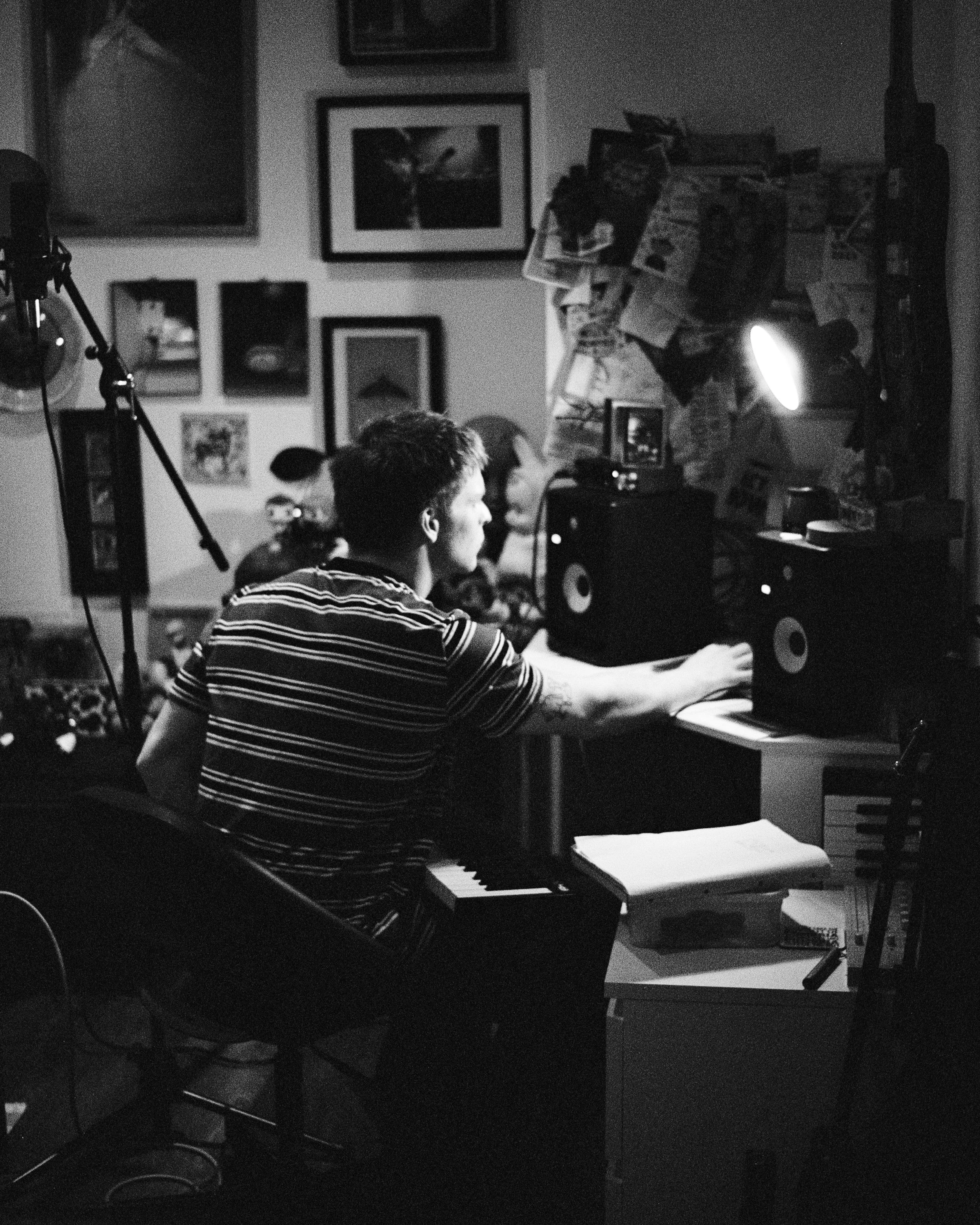 A black-and-white photo of a young man playing a keyboard in a room decorated with framed pictures on the wall. There are studio lights and audio equipment around him.