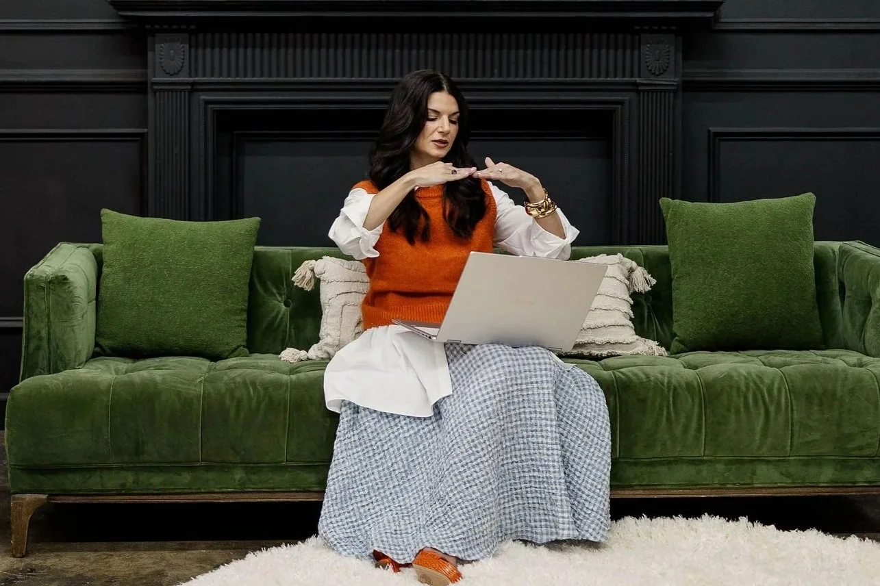 Woman sitting on a green velvet sofa with green pillows, working on a laptop, in a room with a black fireplace mantel and dark wall paneling.