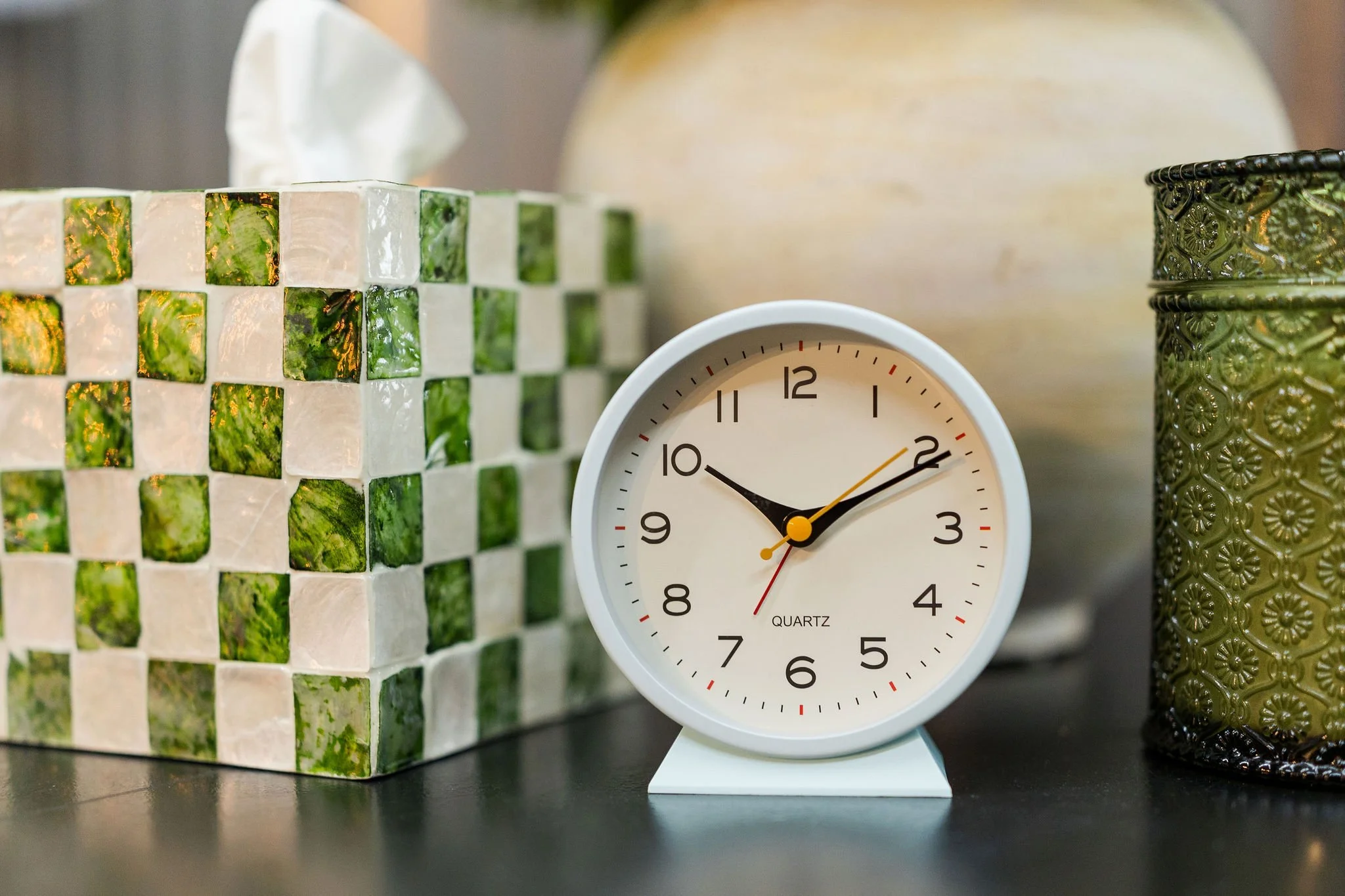 A white quartz clock showing the time as approximately 9:10, placed on a black surface beside a green and beige mosaic tile container and a green decorative metal container.
