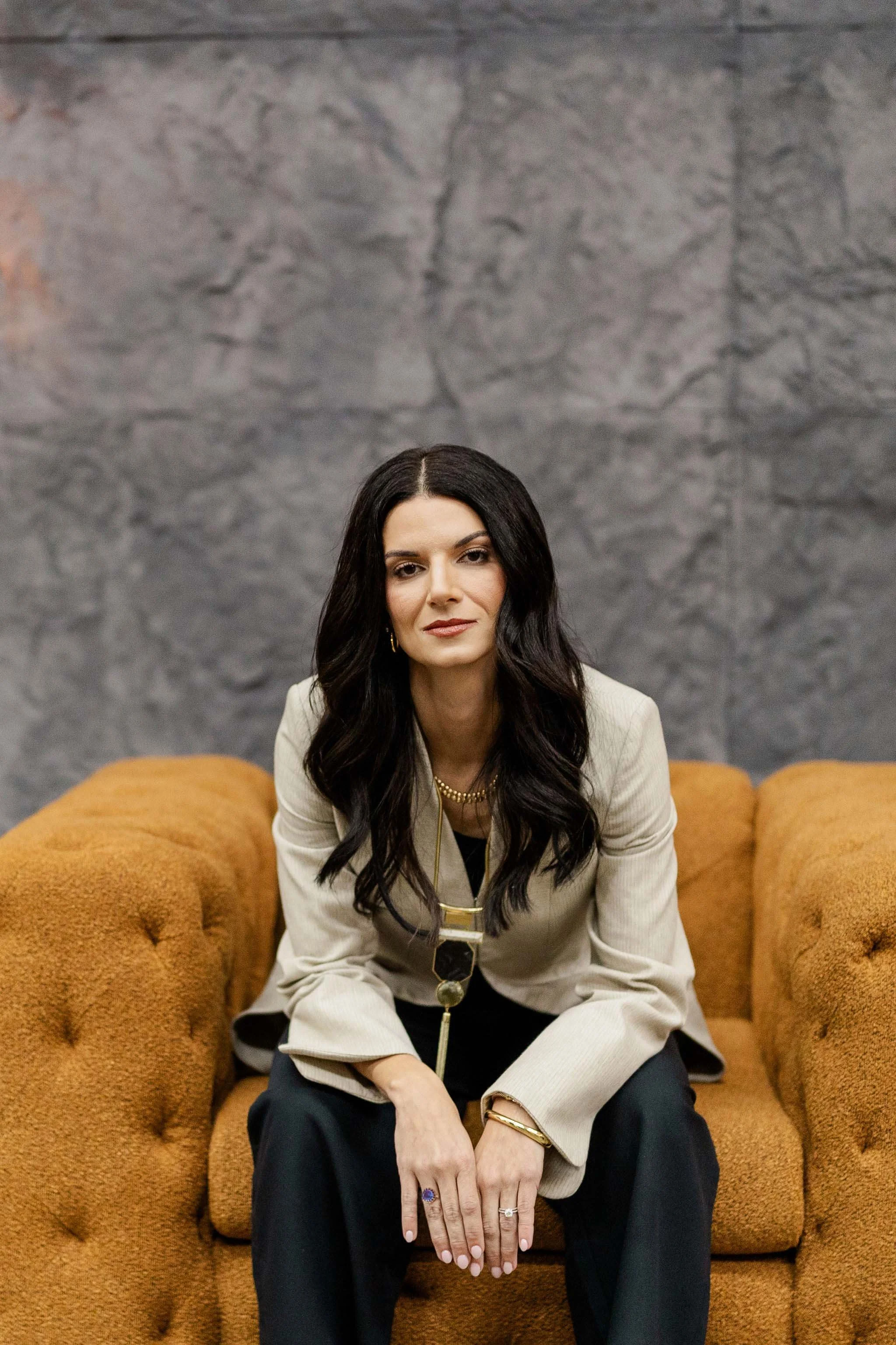 A woman with long dark hair and a beige blazer sitting on a mustard-colored couch, with a gray textured wall in the background.