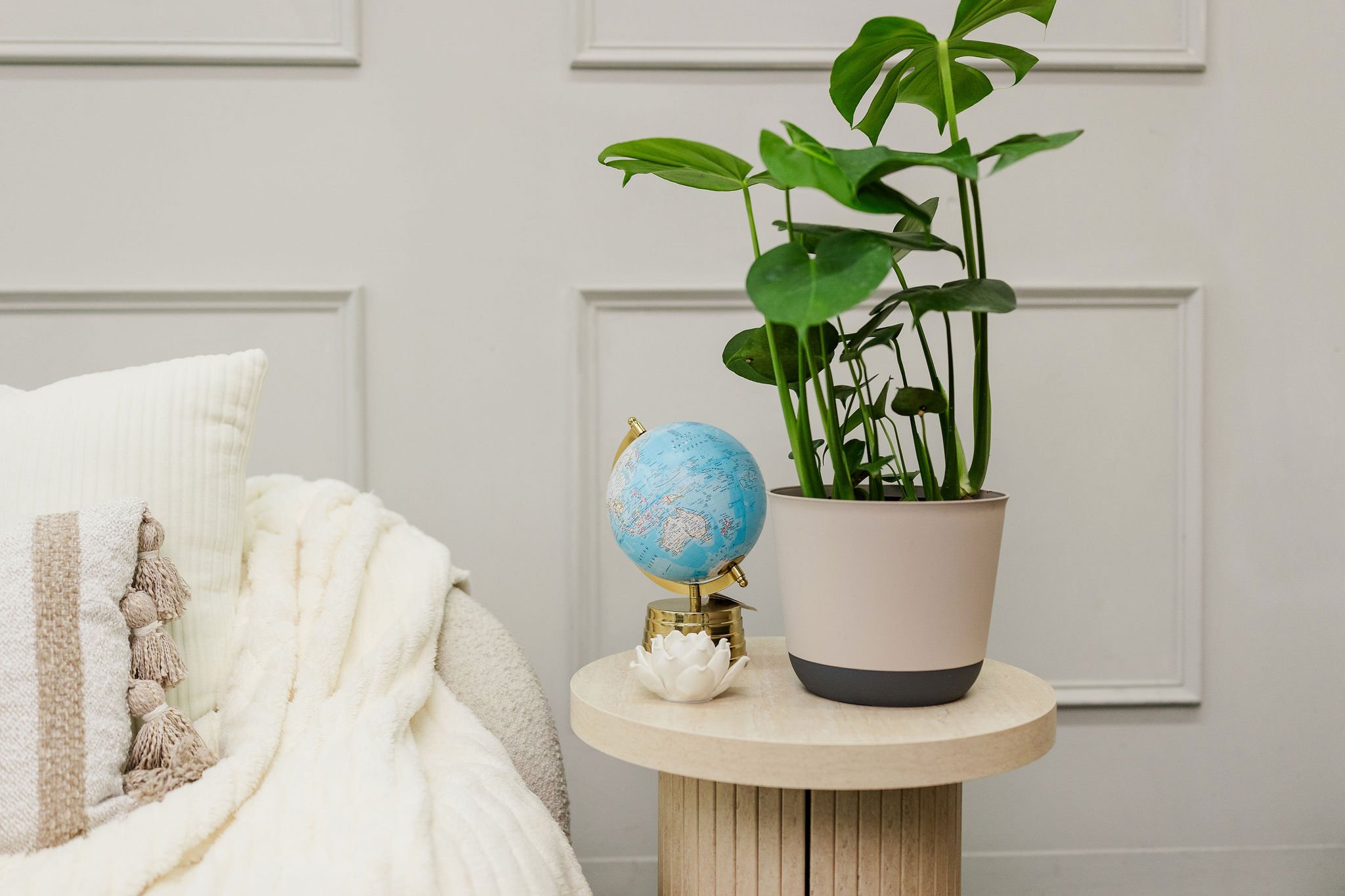 A cozy interior scene featuring a side table with a small globe, a decorative white flower, and a potted monstera plant, next to a white upholstered sofa with a textured throw blanket.