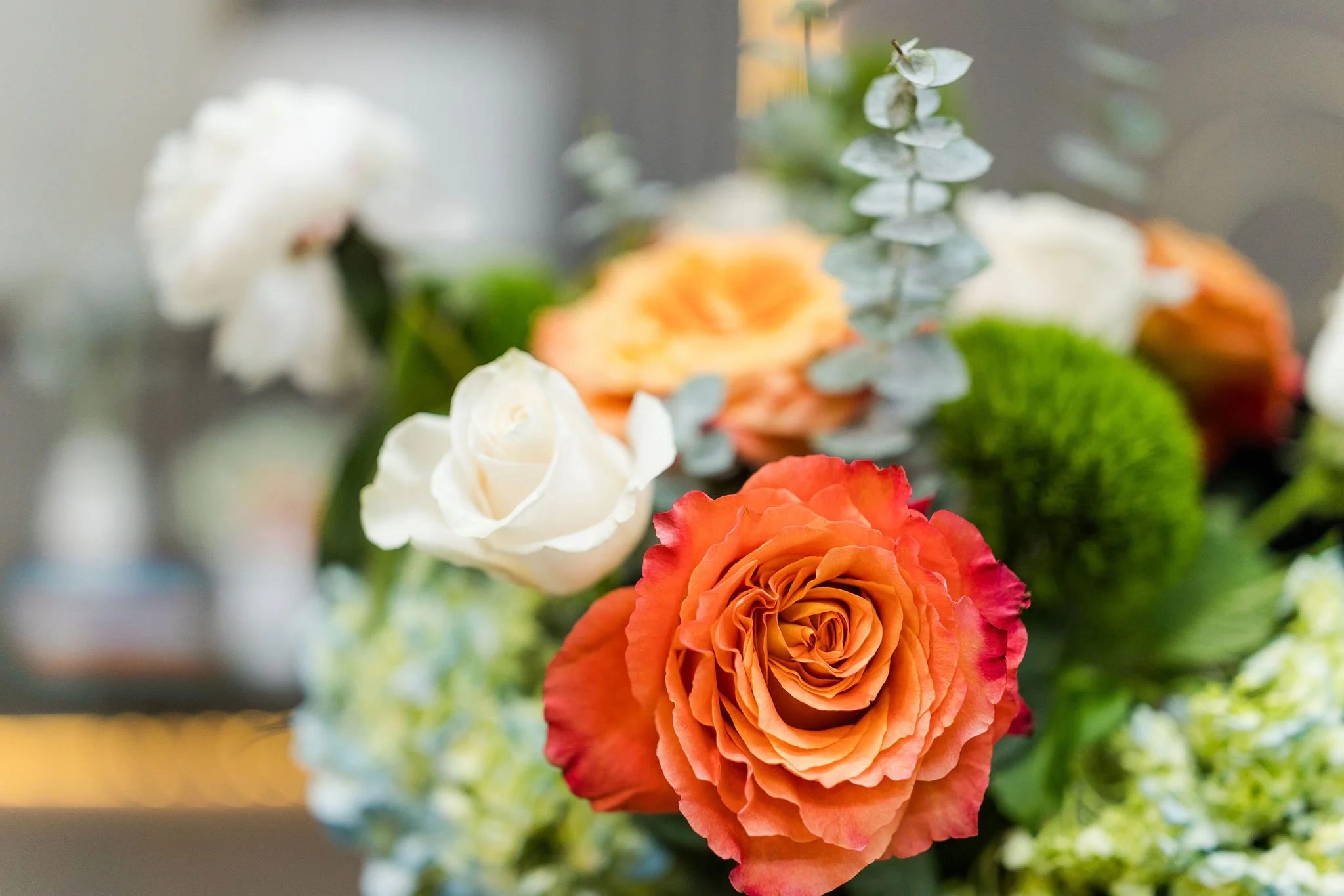 Close-up of a vibrant orange rose with ruffled petals in the foreground, blurred white, cream, pink, and green flowers in the background.