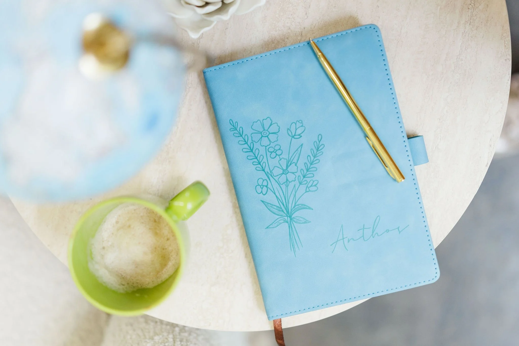 A blue notebook with a floral design on the cover and the name 'Anthos' written on it, a gold pen resting on top, a partially visible mug with a green interior, and a blurred vase of white flowers on a light-colored table.