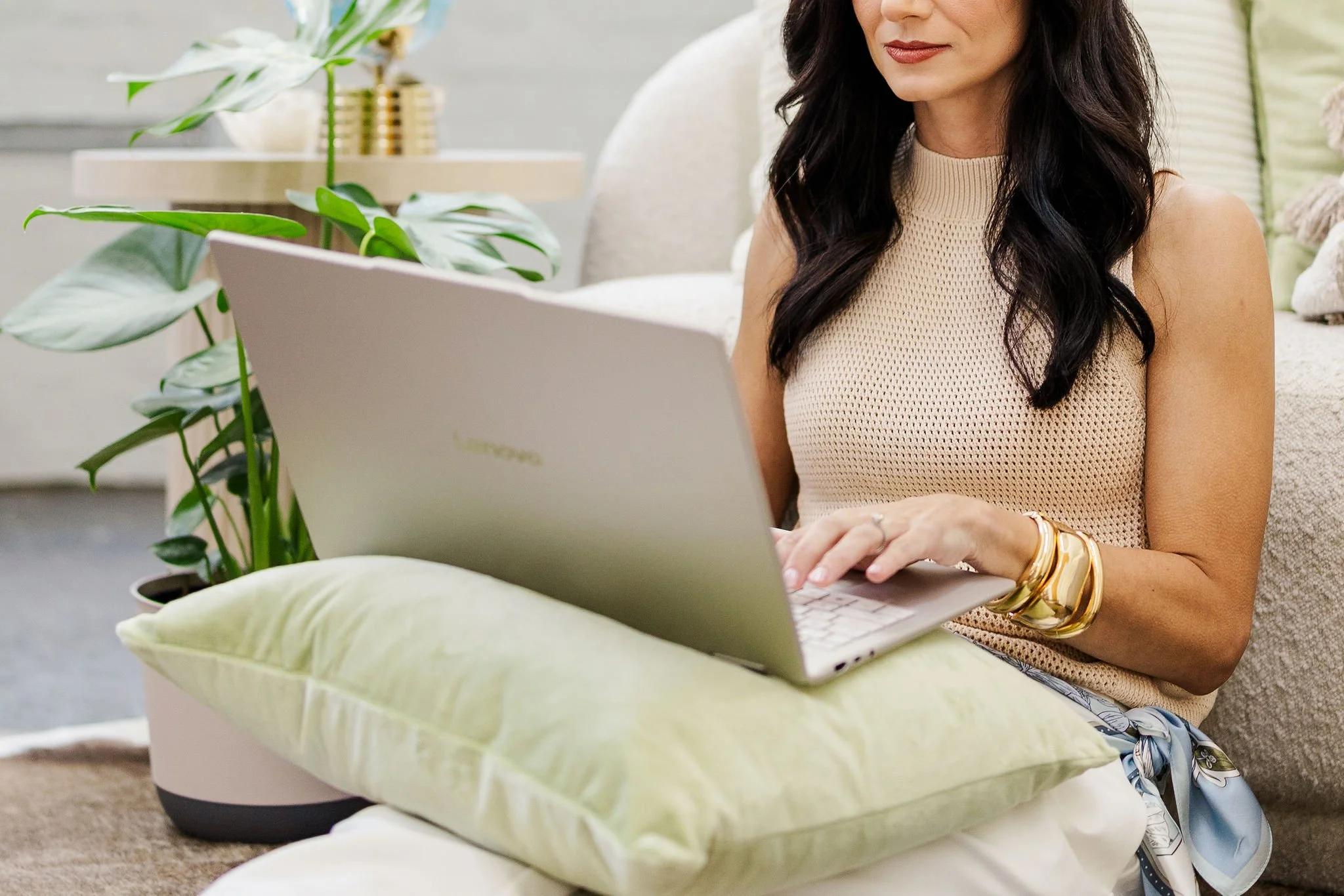 Woman with dark, curled hair sitting with a silver laptop in her lap on top of a green pillow and potted plant nearby in a cozy, well-lit therapy office.