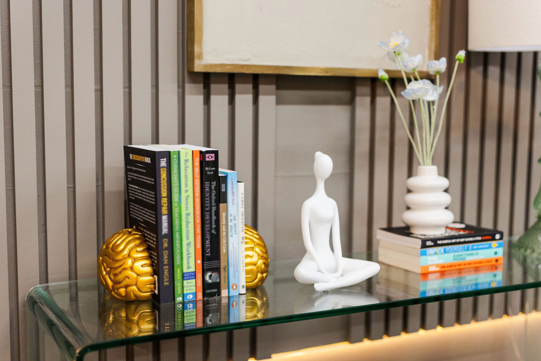 Books on a glass table with decorative gold brain-shaped objects, a white abstract human figure, and a white vase with white flowers, against a background of wooden slats and a framed empty canvas.