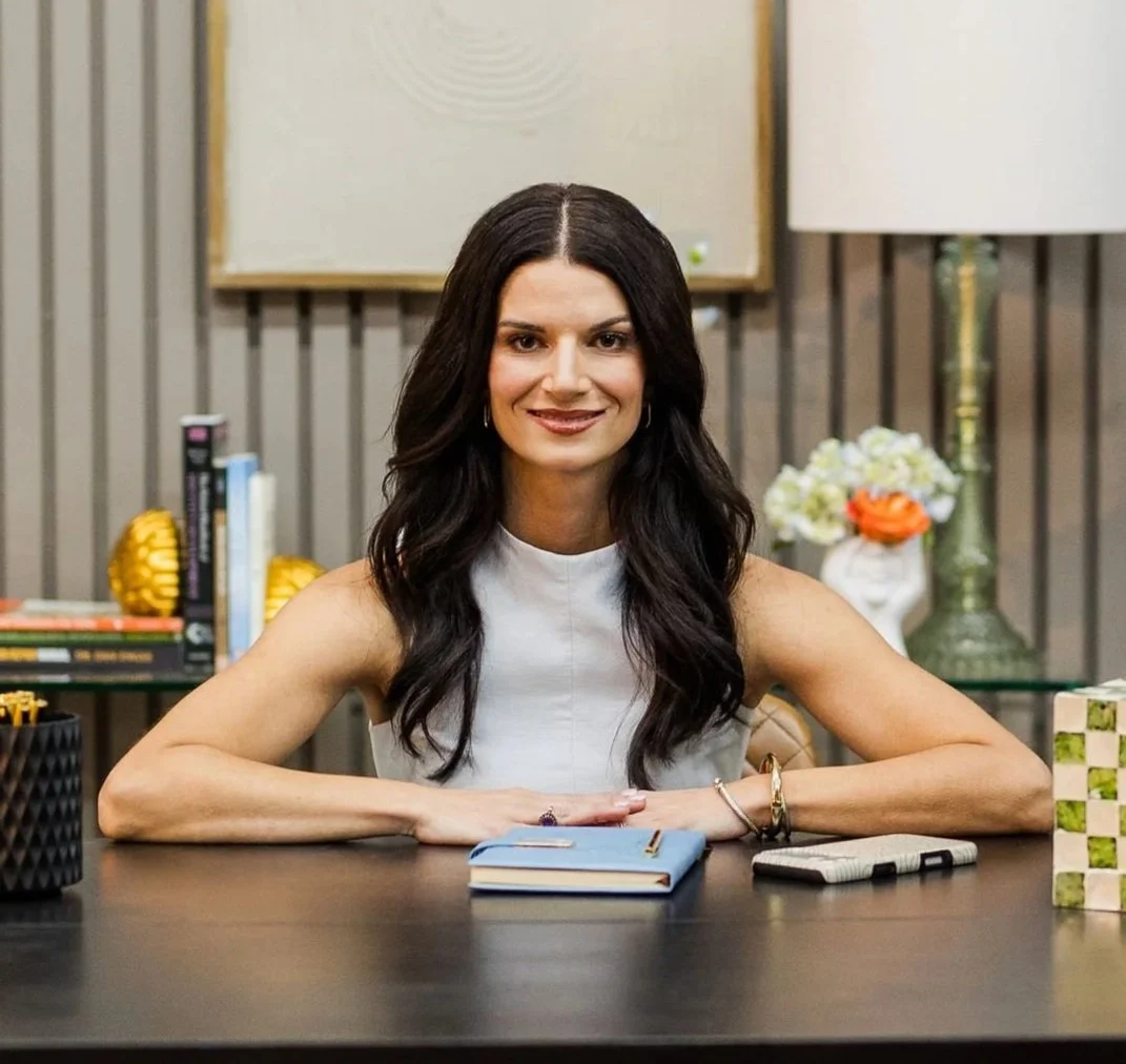 Anna Koullas, a female therapist with long dark hair sitting at a desk, smiling, in a therapy office with books, decorative items, and artwork in the background.