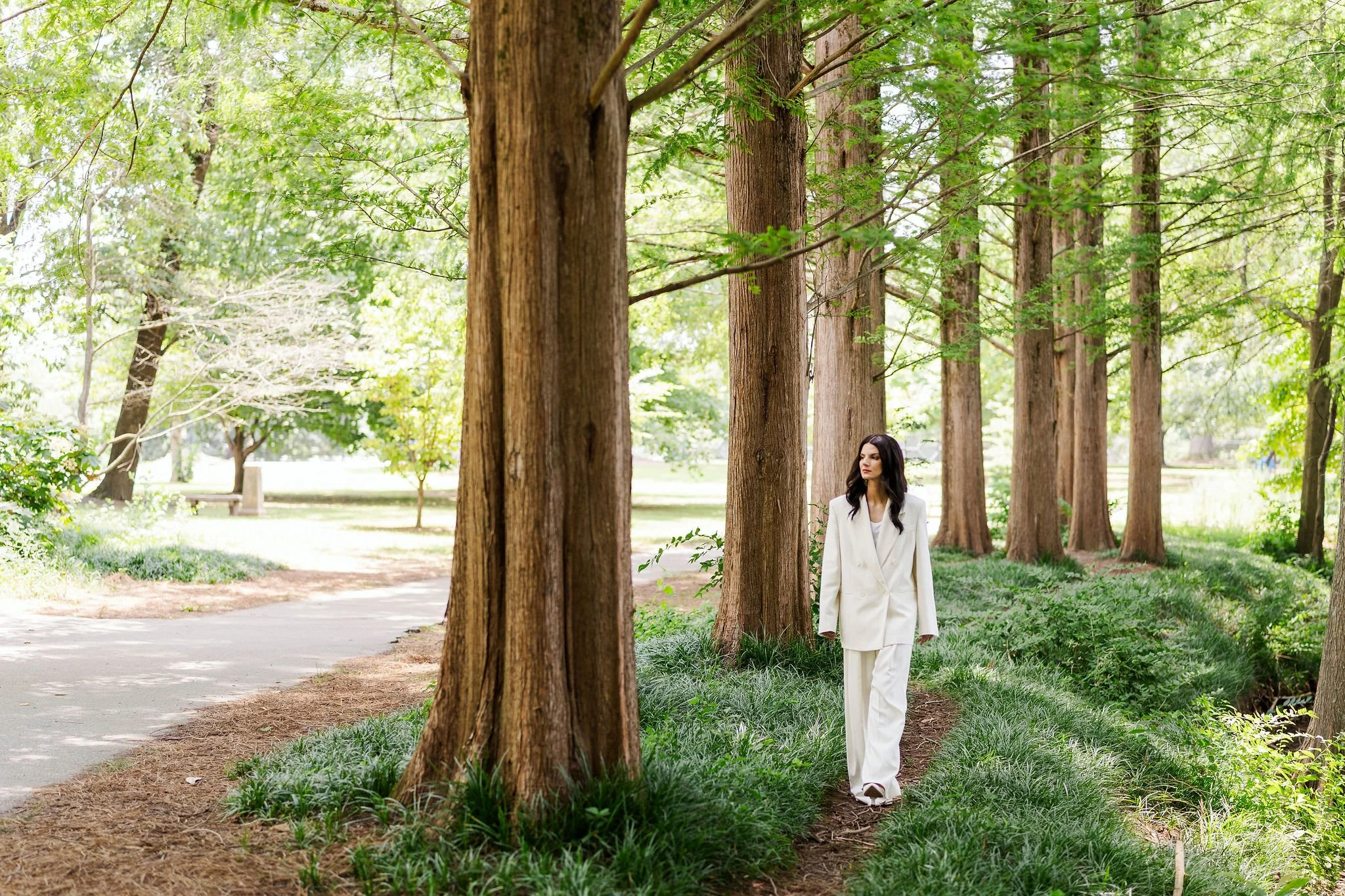 Anna Koullas, a systemic therapist in Atlanta, GA. Anna has long dark hair and is wearing a white suit walking through a park with tall trees and green grass