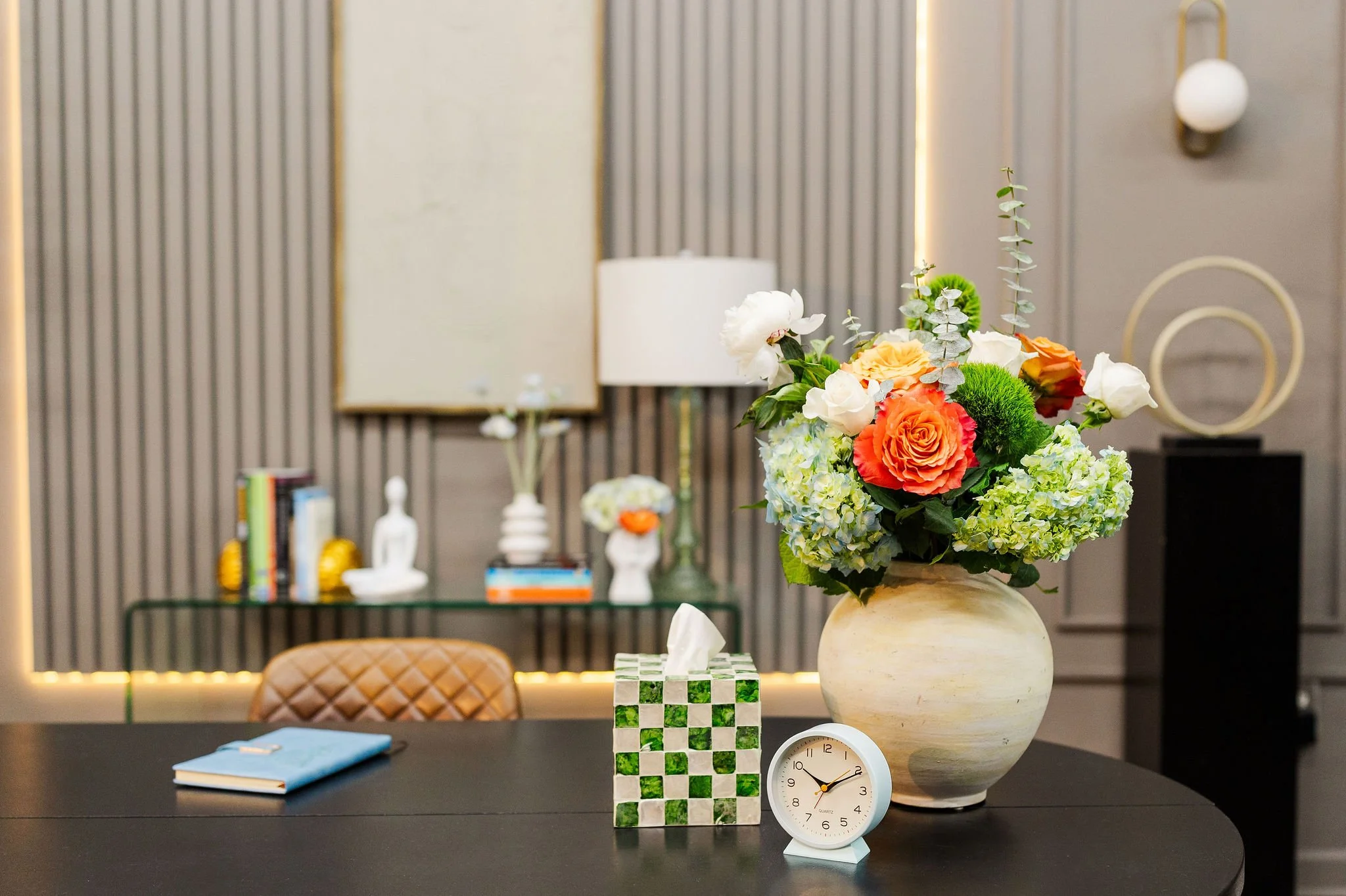 A bouquet of flowers in a beige vase, placed on a black table in a modern room with striped gray and beige walls, with a decorative lamp, books, and a clock in the background.