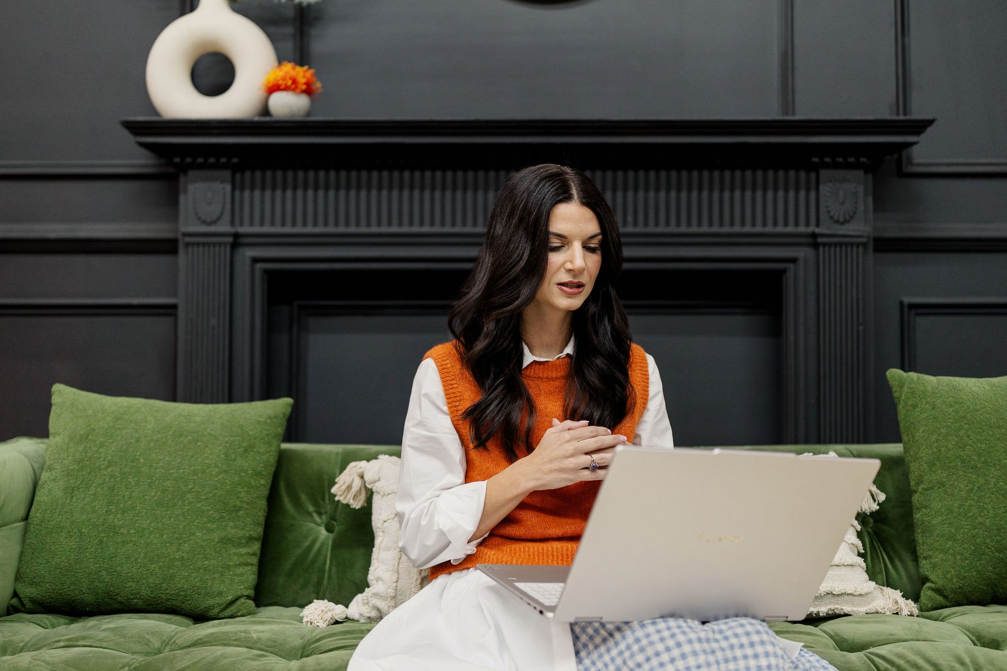 A female therapist with long dark hair, wearing an orange vest over a white shirt, is sitting on a green sofa in a room with dark gray walls and a black mantle. She is looking at her laptop while talking to a therapy patient during a virtual session.