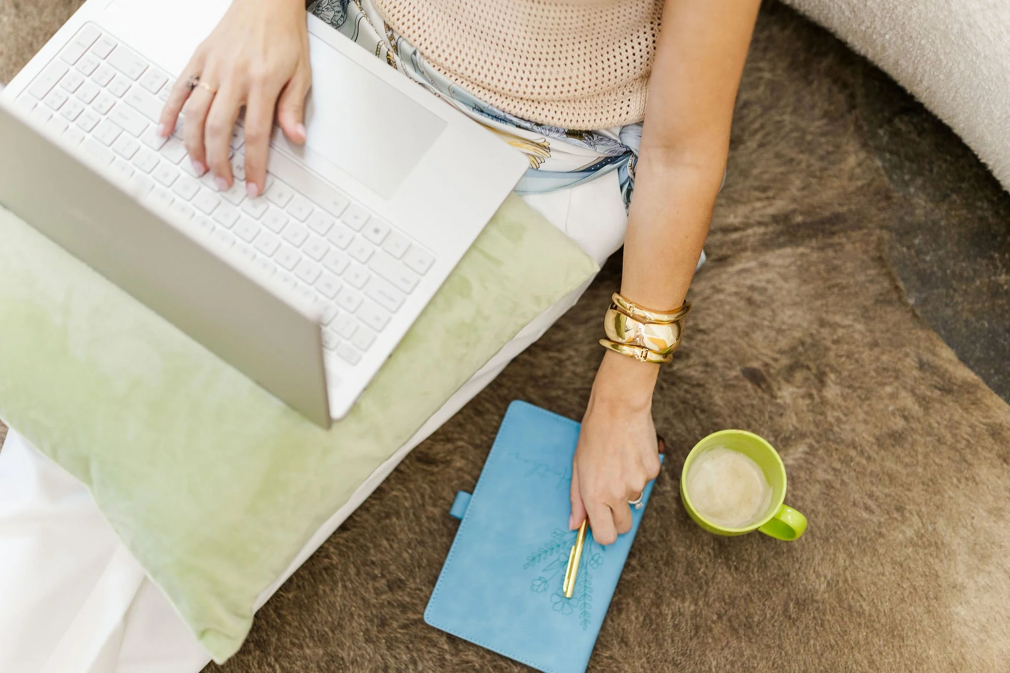 Person sitting on brown carpeted floor, reaching for a blue notebook and gold pen, next to a green mug with a beverage. A laptop is positioned in her lap on top of a light green pillow.