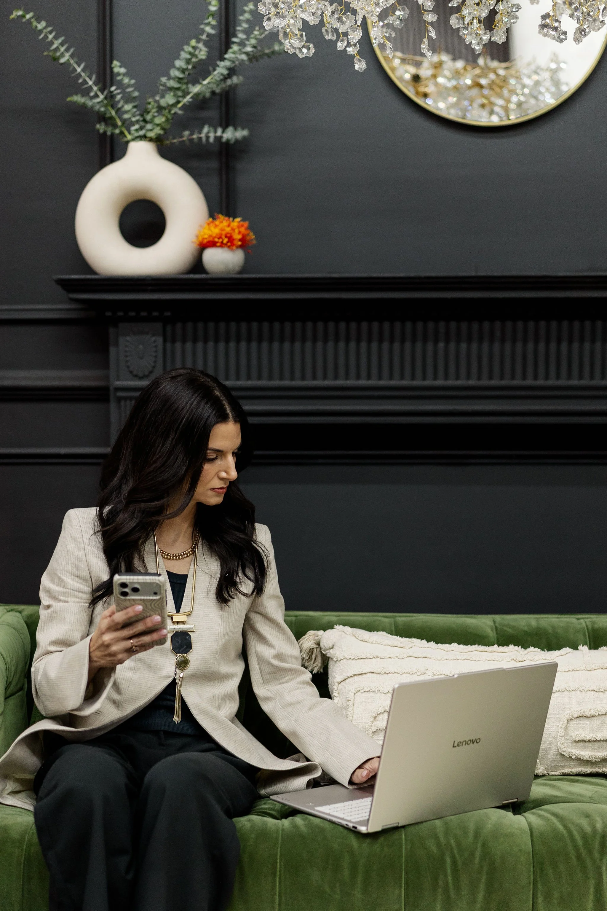 A woman with long dark hair, wearing a beige blazer and black pants, is sitting on a green velvet sofa. She is holding a smartphone in her right hand and has a laptop on her lap. There is a cream-colored blanket on the sofa next to her. In the background, a black fireplace mantel has decorative vases and a circular mirror, with a floral arrangement and a chandelier above.