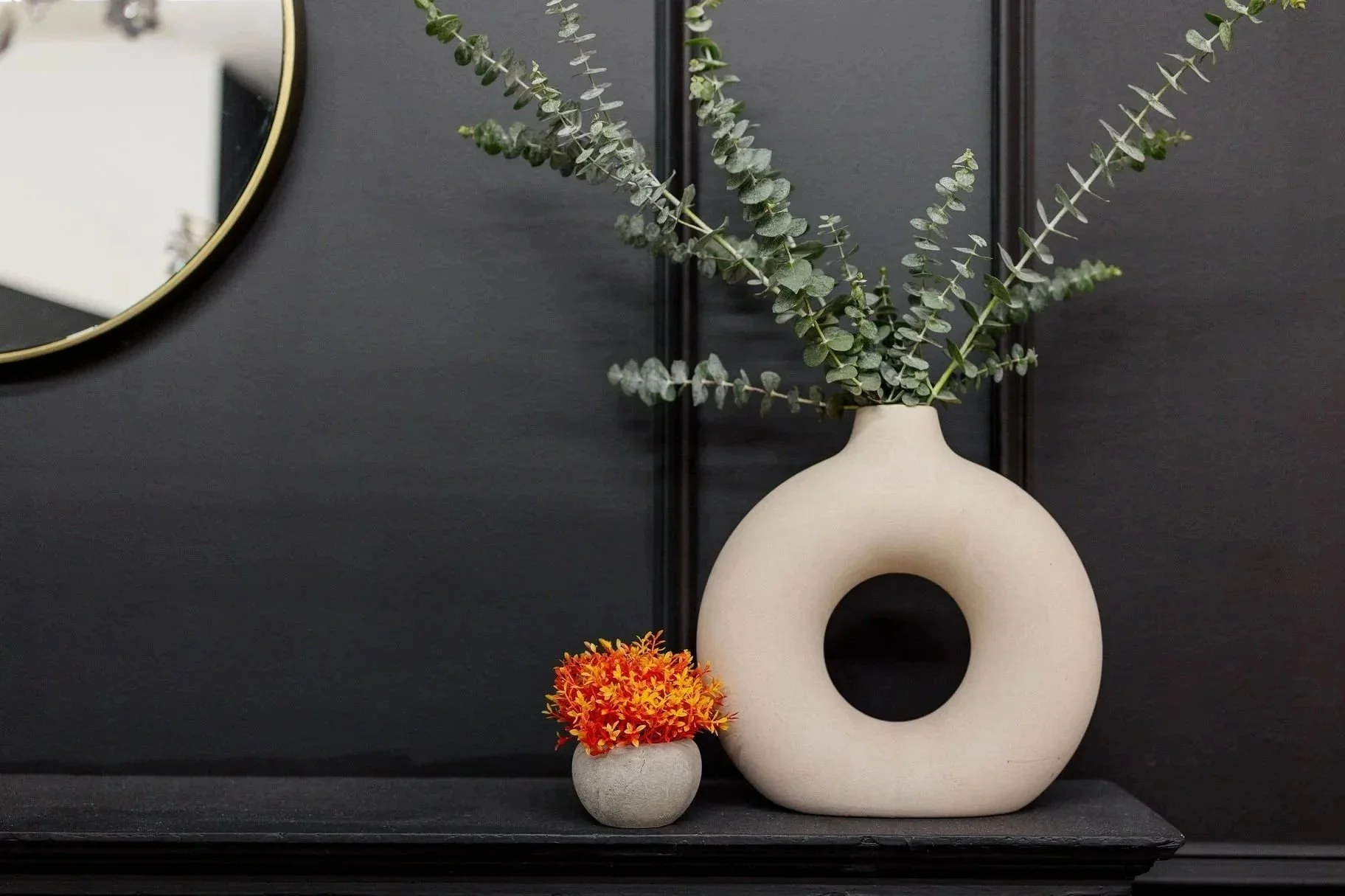 Decorative black wall with a round mirror and modern white sculpture vase with eucalyptus branches, a small gray pot with orange flowers, and a black tray.