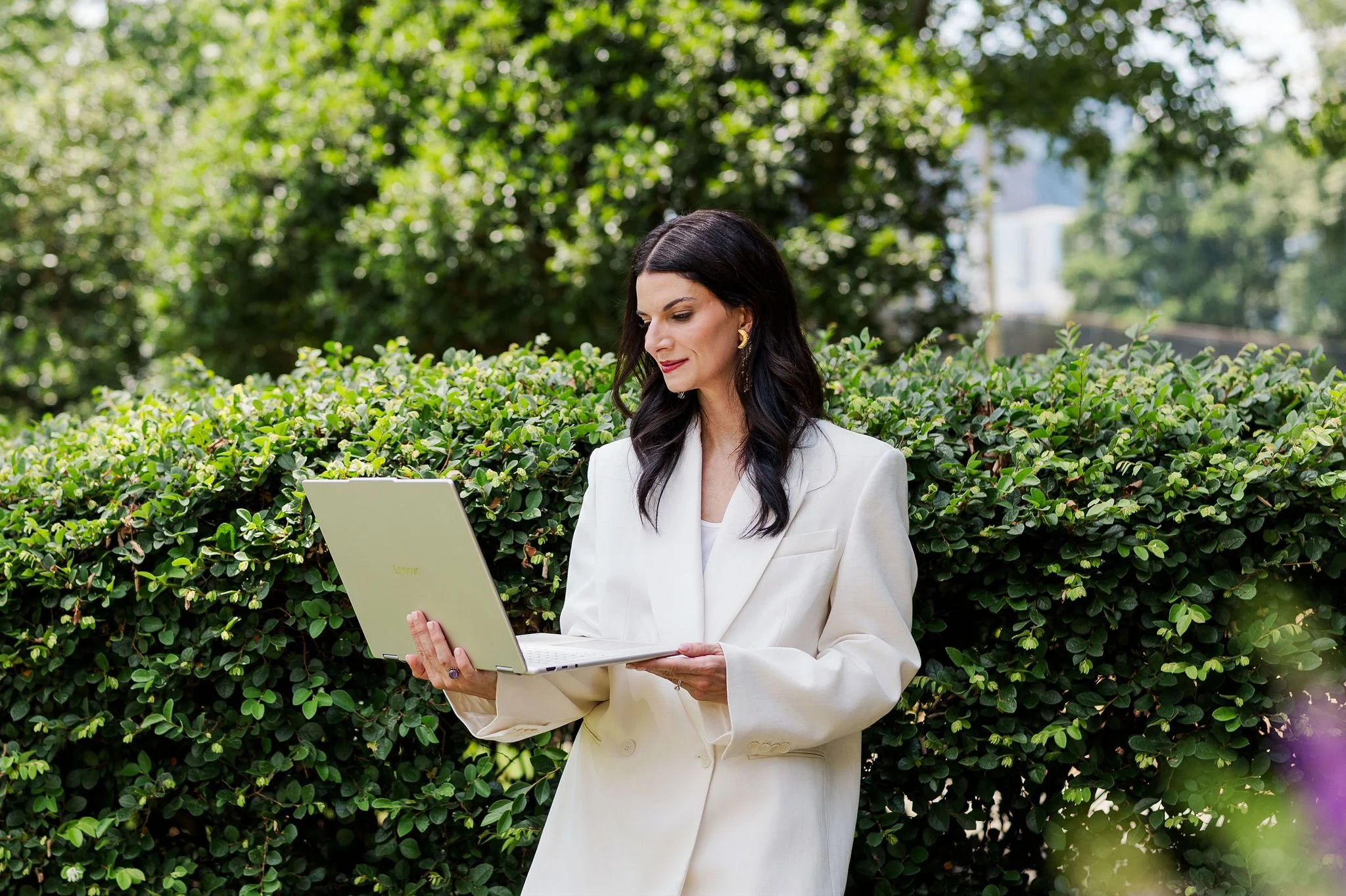 A woman in a white suit standing outdoors and reading a light green laptop in front of a green hedge.