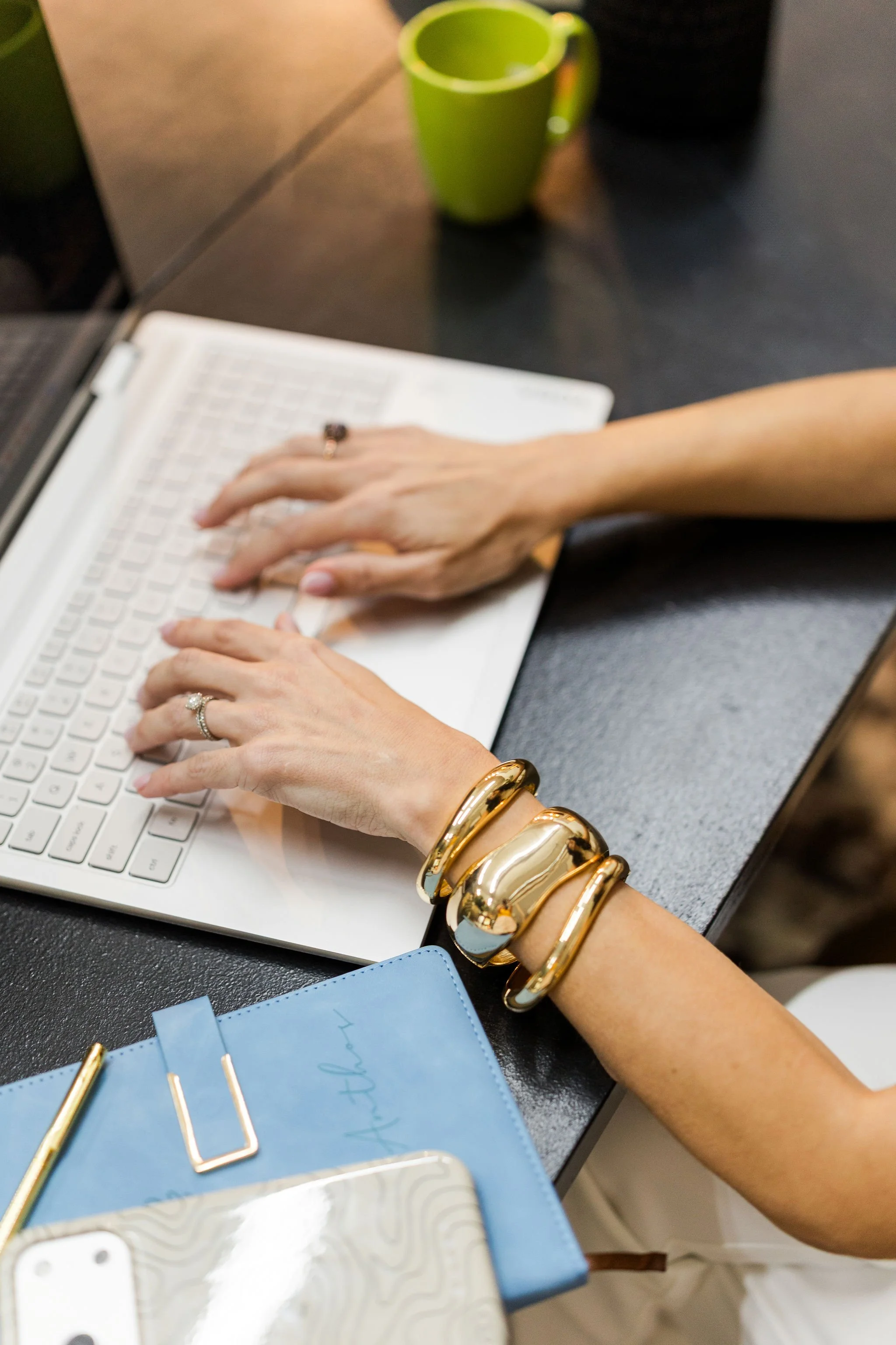 A person wearing multiple gold bangles, rings, and a watch is typing on a white keyboard. There are green and black cups, a blue planner, gold paper clips, and a white phone on the desk.