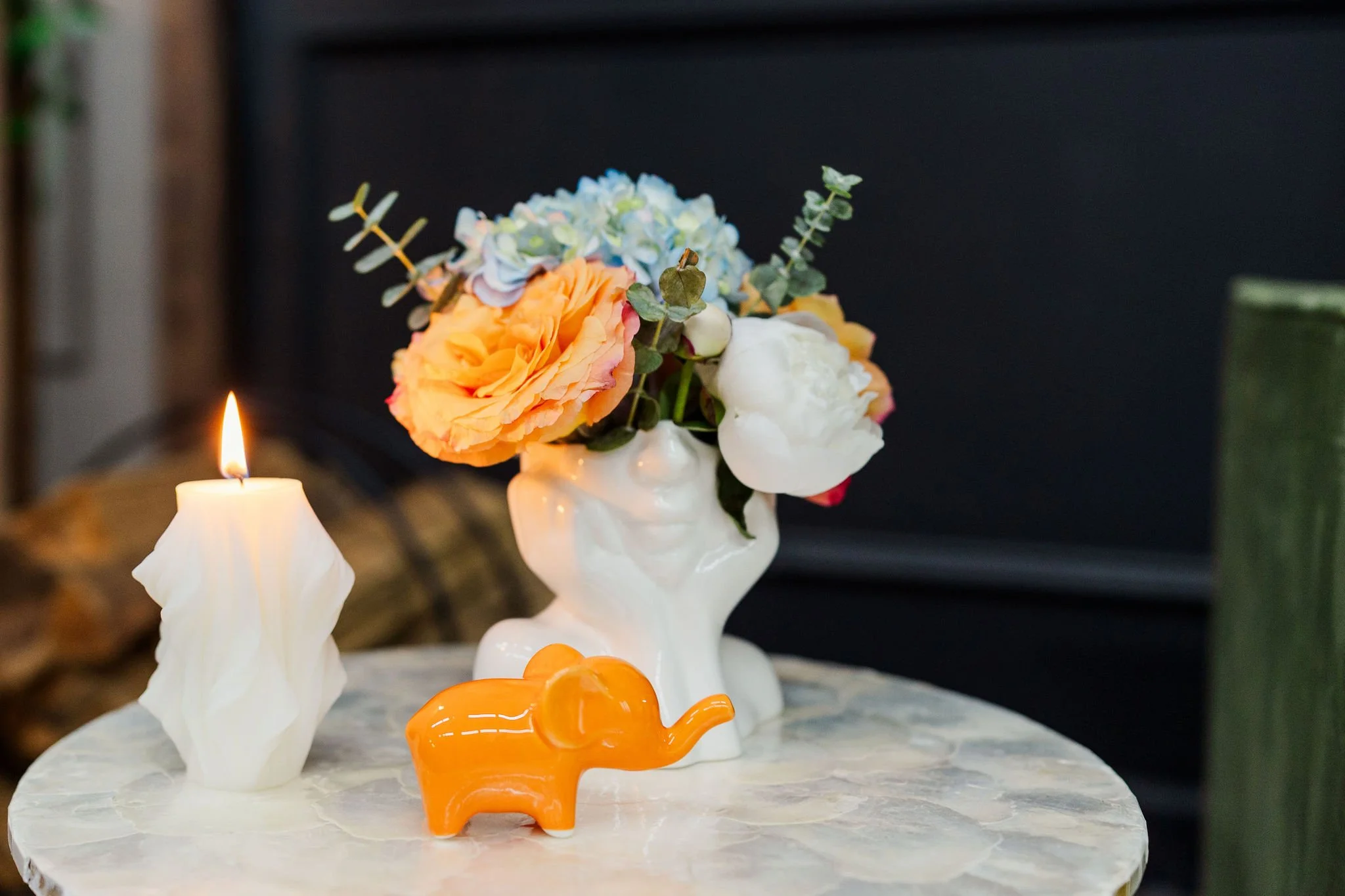 A floral arrangement in a white vase with orange, white, and blue flowers, next to a lit candle, small orange elephant figurine, and decorative white and orange objects on a marble surface.