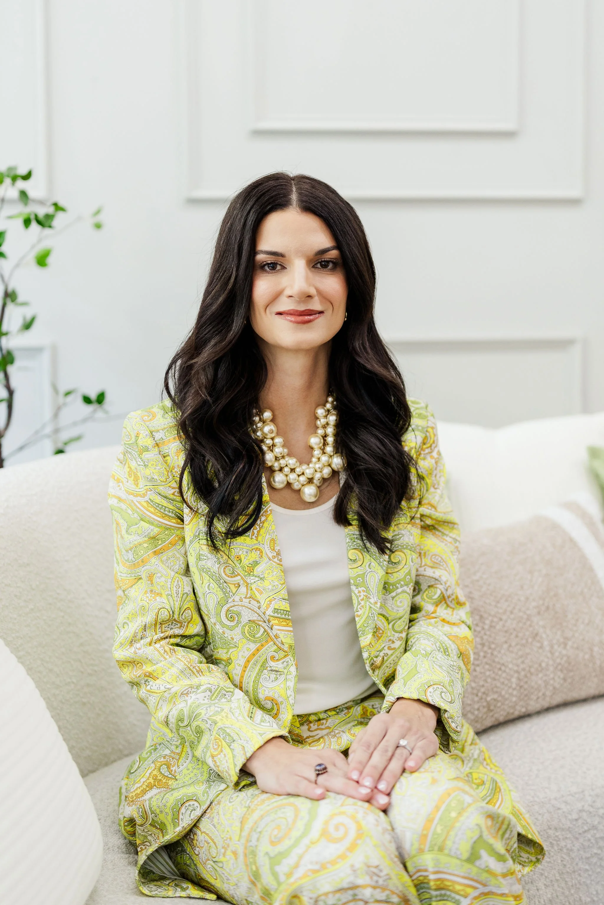 A woman with long dark hair, wearing a yellow paisley-patterned blazer and pants, a white top, and a pearl necklace, sitting on a white chair in a bright room with white walls and a green plant in the background.