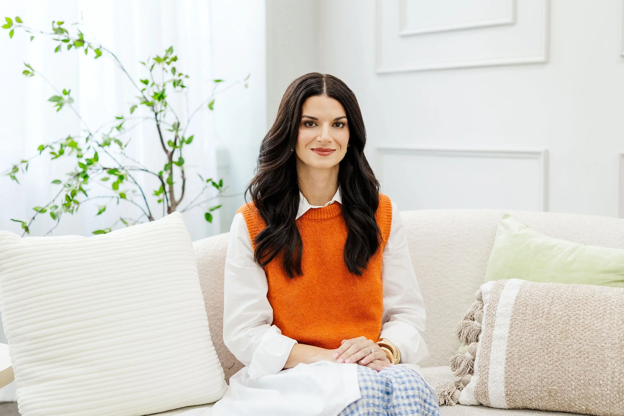 Anna Koullas, a systemic therapist with long dark hair, wearing an orange sweater over a white shirt, sitting on a light-colored sofa in a bright, therapy office with white walls, green plants, and cushions.