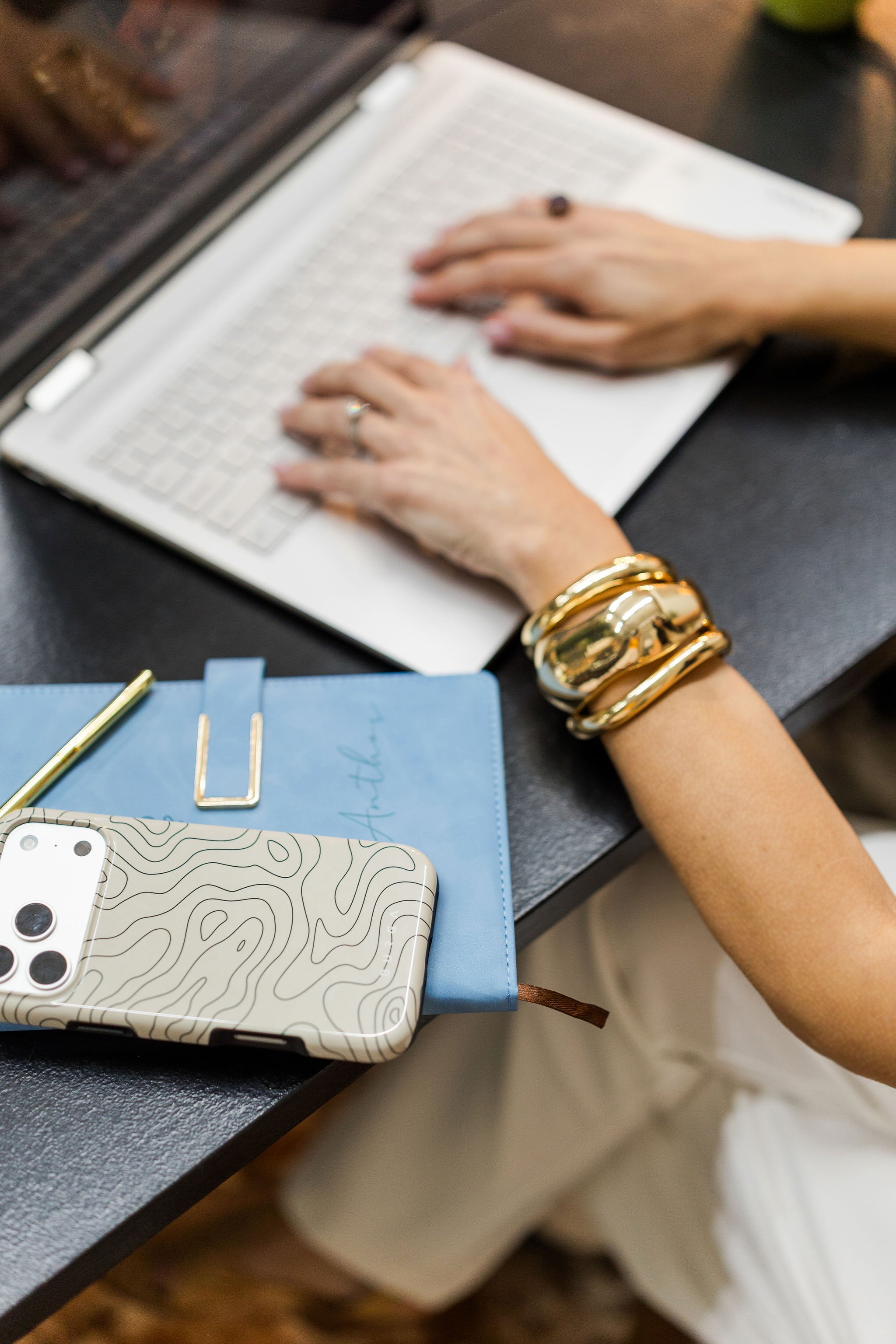 A female therapist with a gold bracelet and rings typing on a white keyboard at a black desk. Nearby are a blue notebook with writing, a smartphone with a patterned case, and a pen.