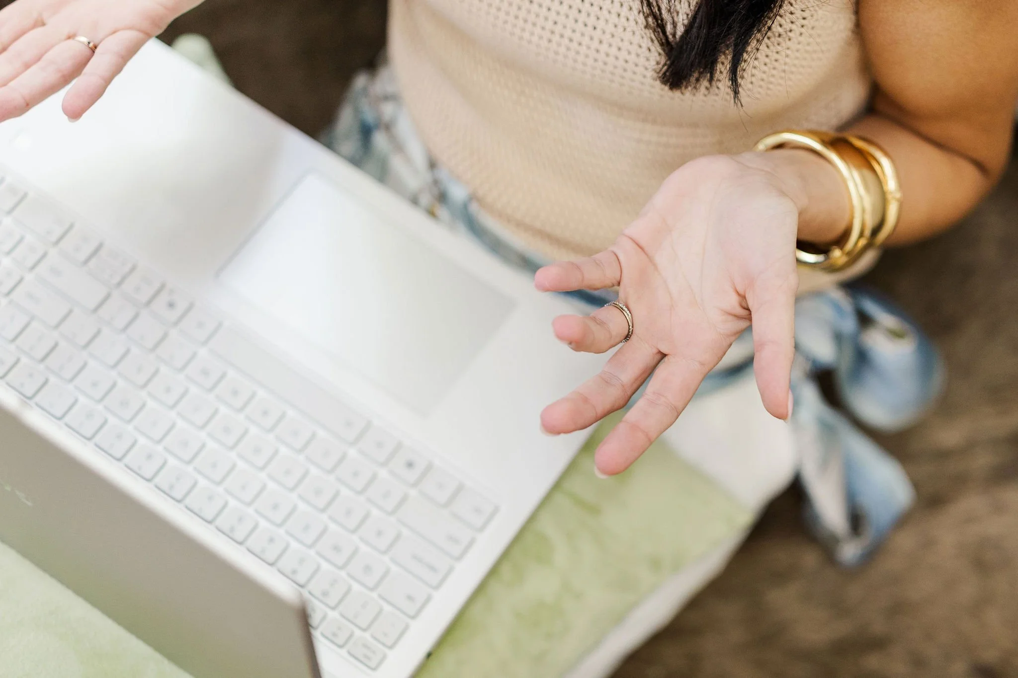 A person using a white laptop while gesturing with their hands, wearing rings and gold bracelets, sitting at a table with a green surface.