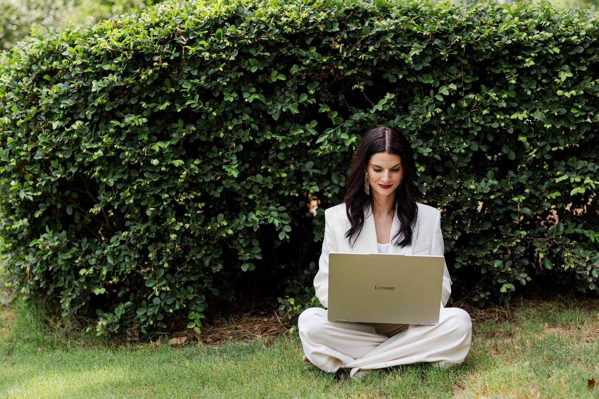 A woman with dark hair and earrings sitting cross-legged on grass in front of a large green bush, working on a silver Lenovo laptop.