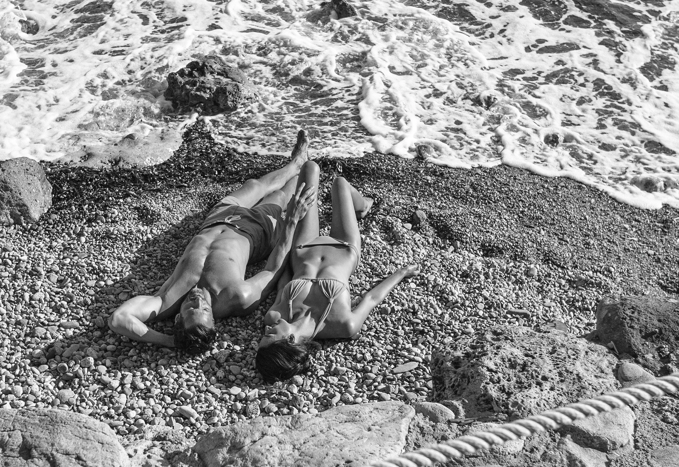 Three people lying on a pebble beach near the shoreline, with waves crashing nearby.