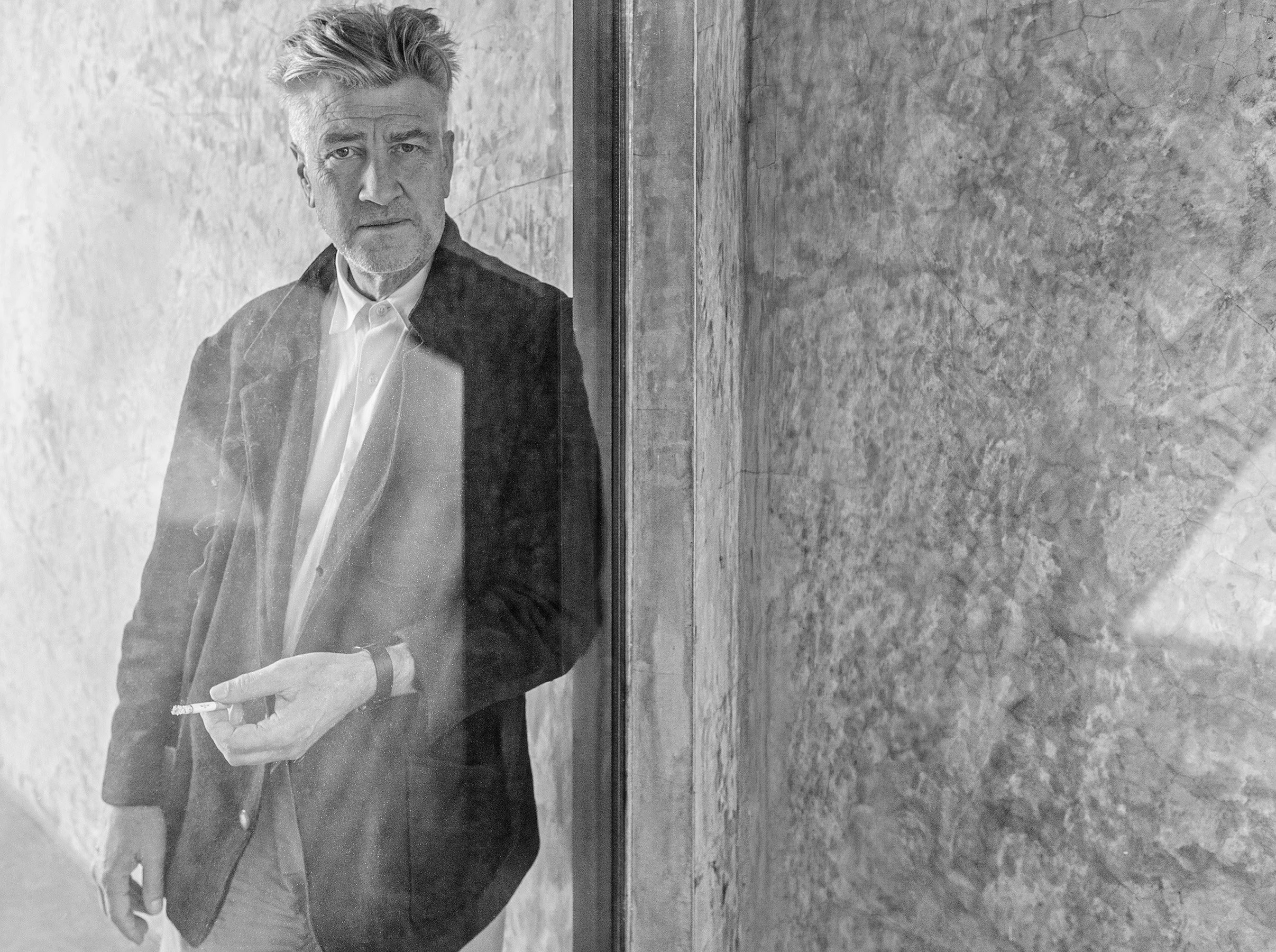 David Lynch, wearing a dark blazer and white shirt, holding a cigarette, and standing in front of a textured wall and a glass window or door.