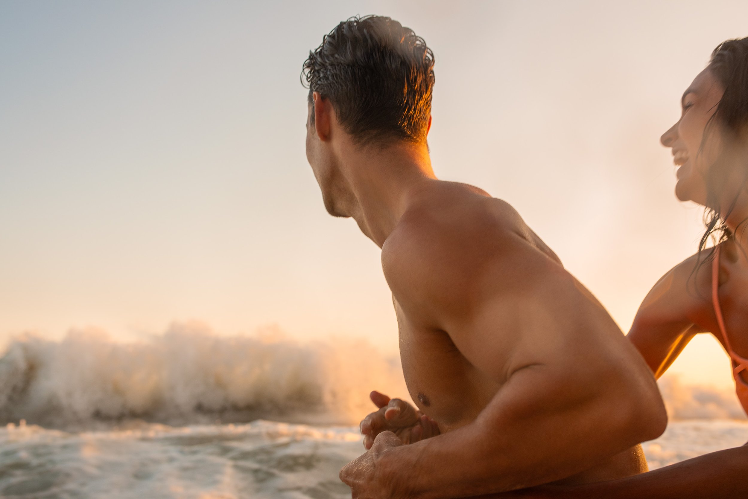 A man and woman practicing yoga or meditation together on the beach during sunset, facing the ocean.