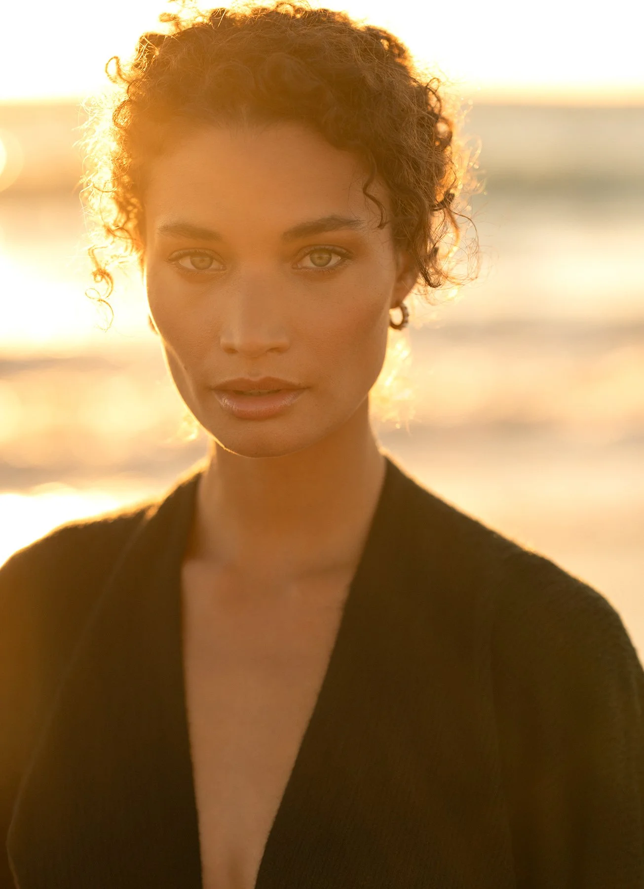 A woman with curly hair and light eyes is shown close-up, facing the camera during sunset at the beach, wearing a black top or coat. Fashion, resort, swimwear, luxury lifestyle photographer. 