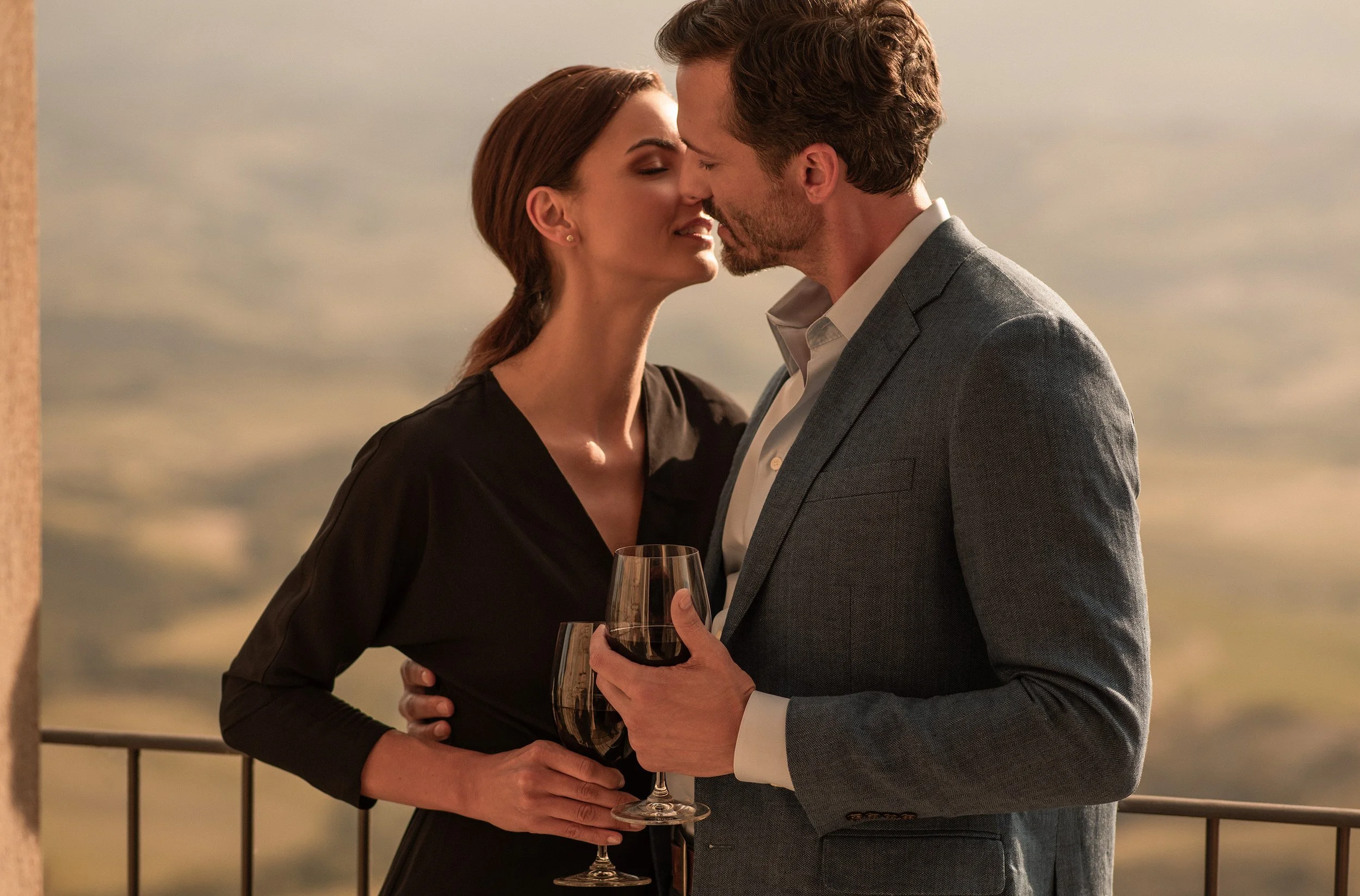 A man and woman dressed in formal attire, sharing an intimate moment with their faces close together against the backdrop of a cloudy sky. The woman holds a glass of red wine, and the man also holds a glass of red wine.