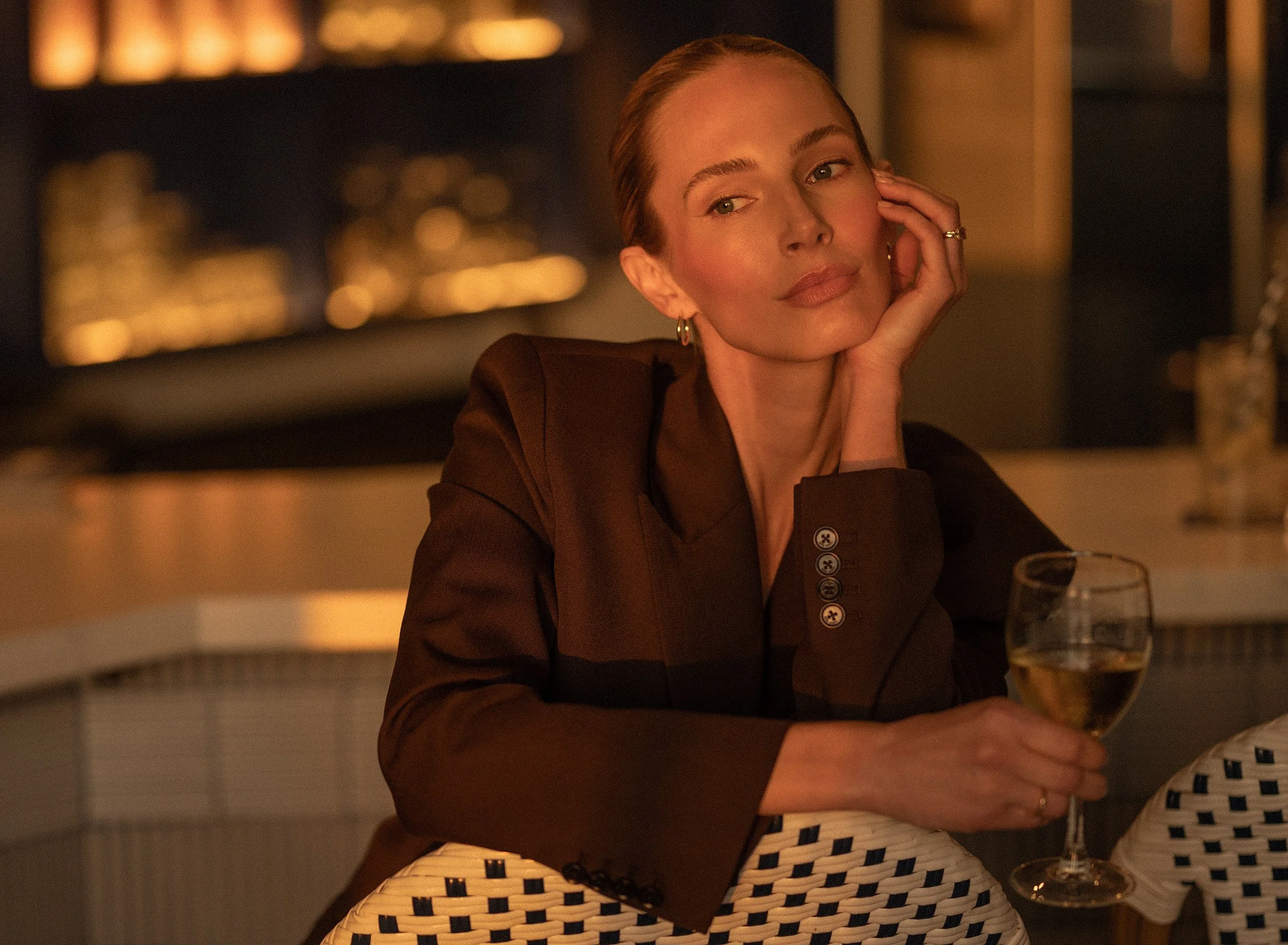A woman with red hair and hoop earrings gazing thoughtfully while resting her head on her hand at a candlelit dinner, with a glass of white wine in front of her.