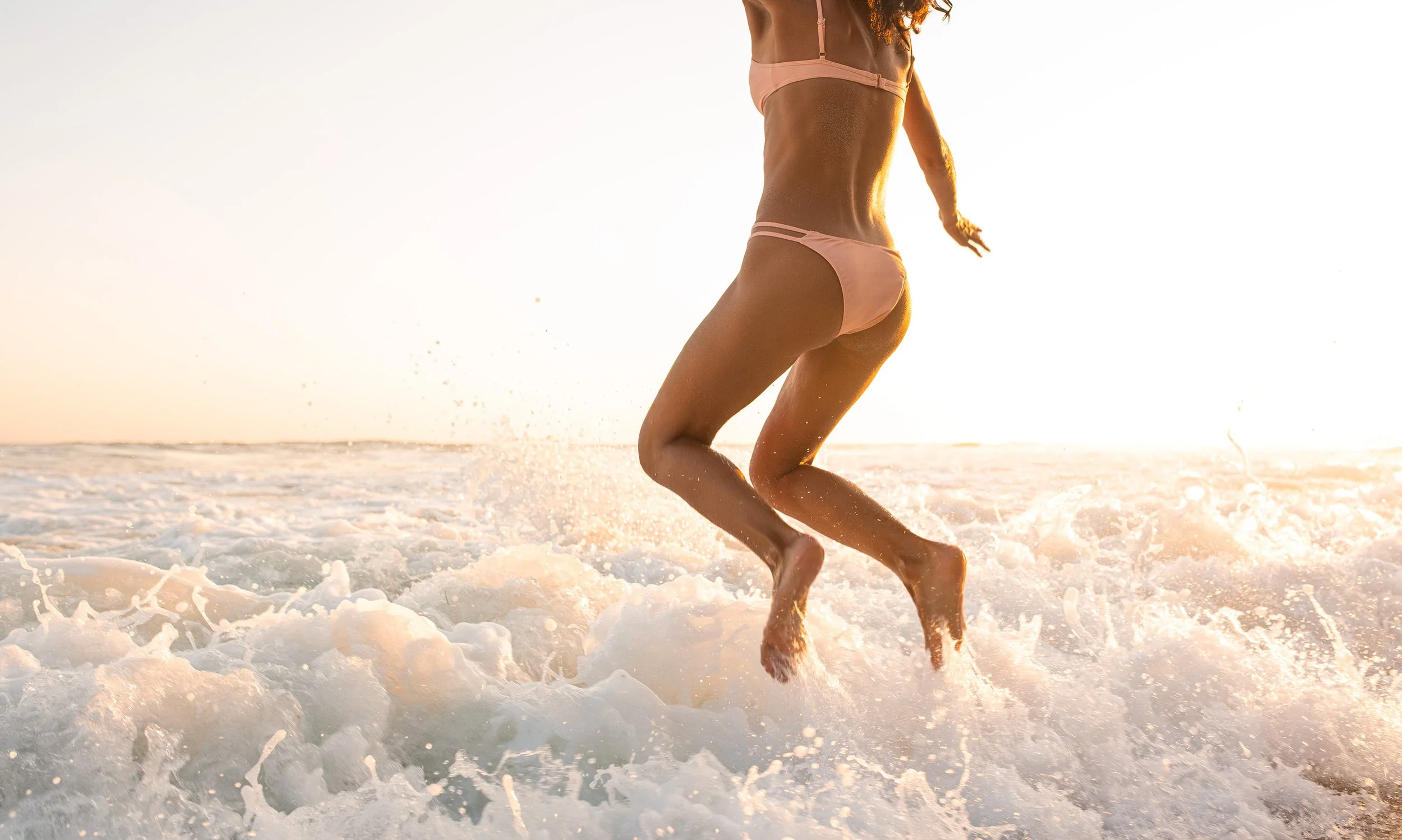 Woman in pink bikini running through shallow ocean water during sunset or sunrise.