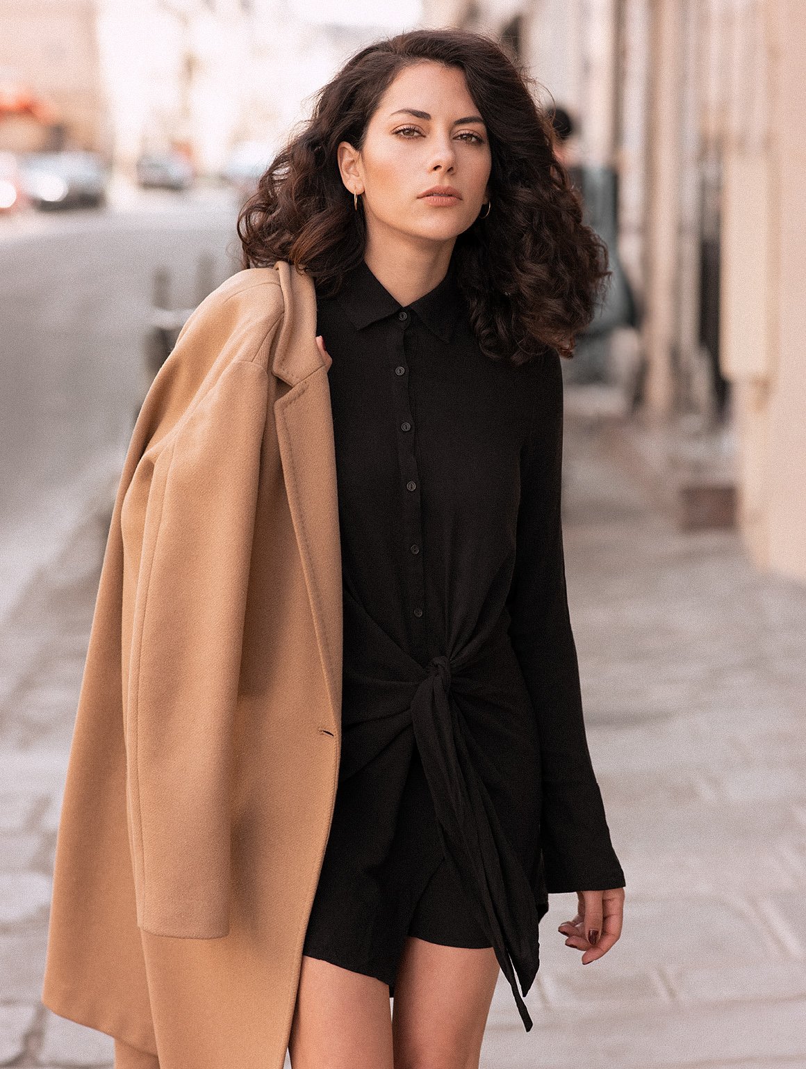 A woman standing on a city sidewalk wearing a black button-up dress with a front tie, carrying a light brown coat over her shoulder, with city buildings and cars in the background.