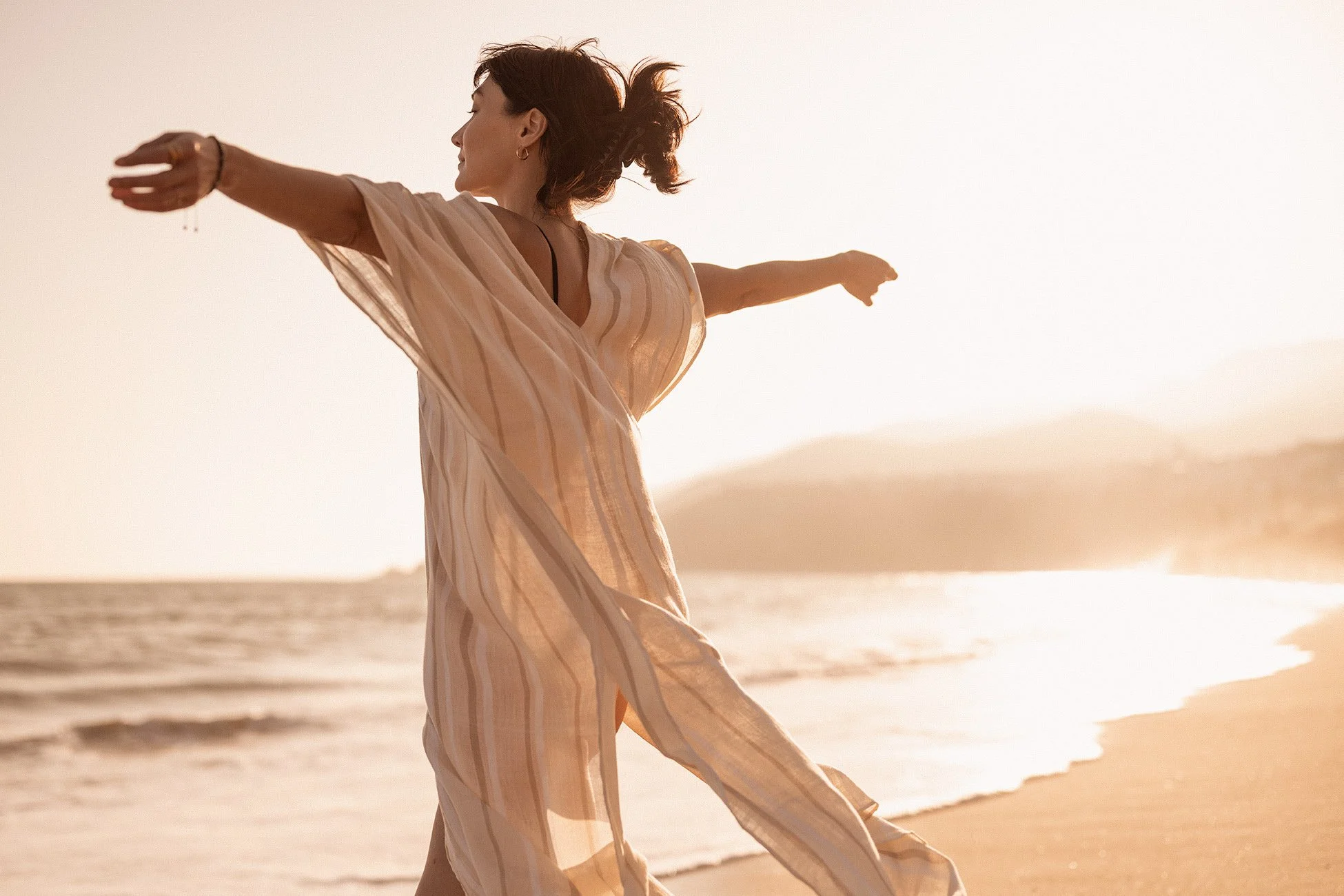 Woman with arms outstretched, wearing a flowing dress, at the beach during sunset.
