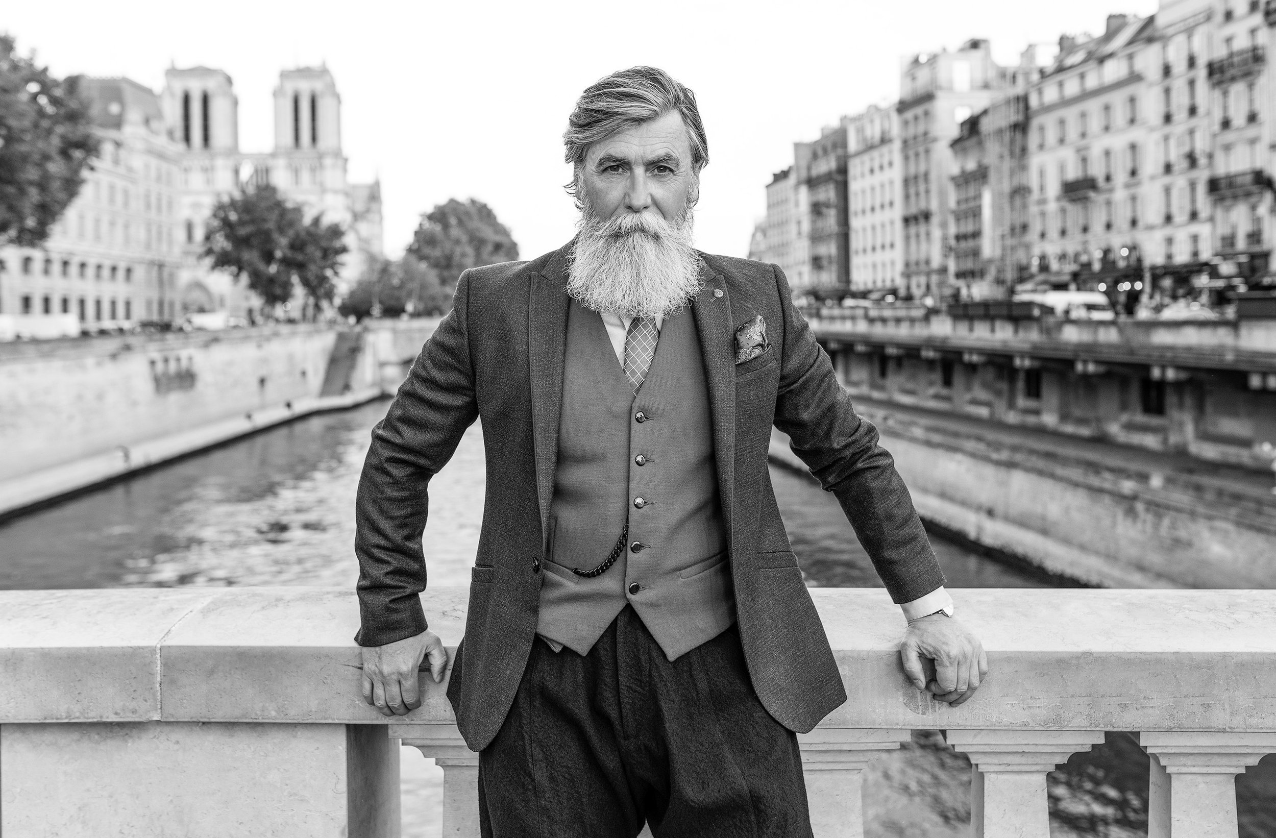 A black and white photo of a distinguished man with a full beard and styled hair, wearing a suit with a vest and tie, standing outdoors on a bridge in front of a cityscape with buildings and trees.