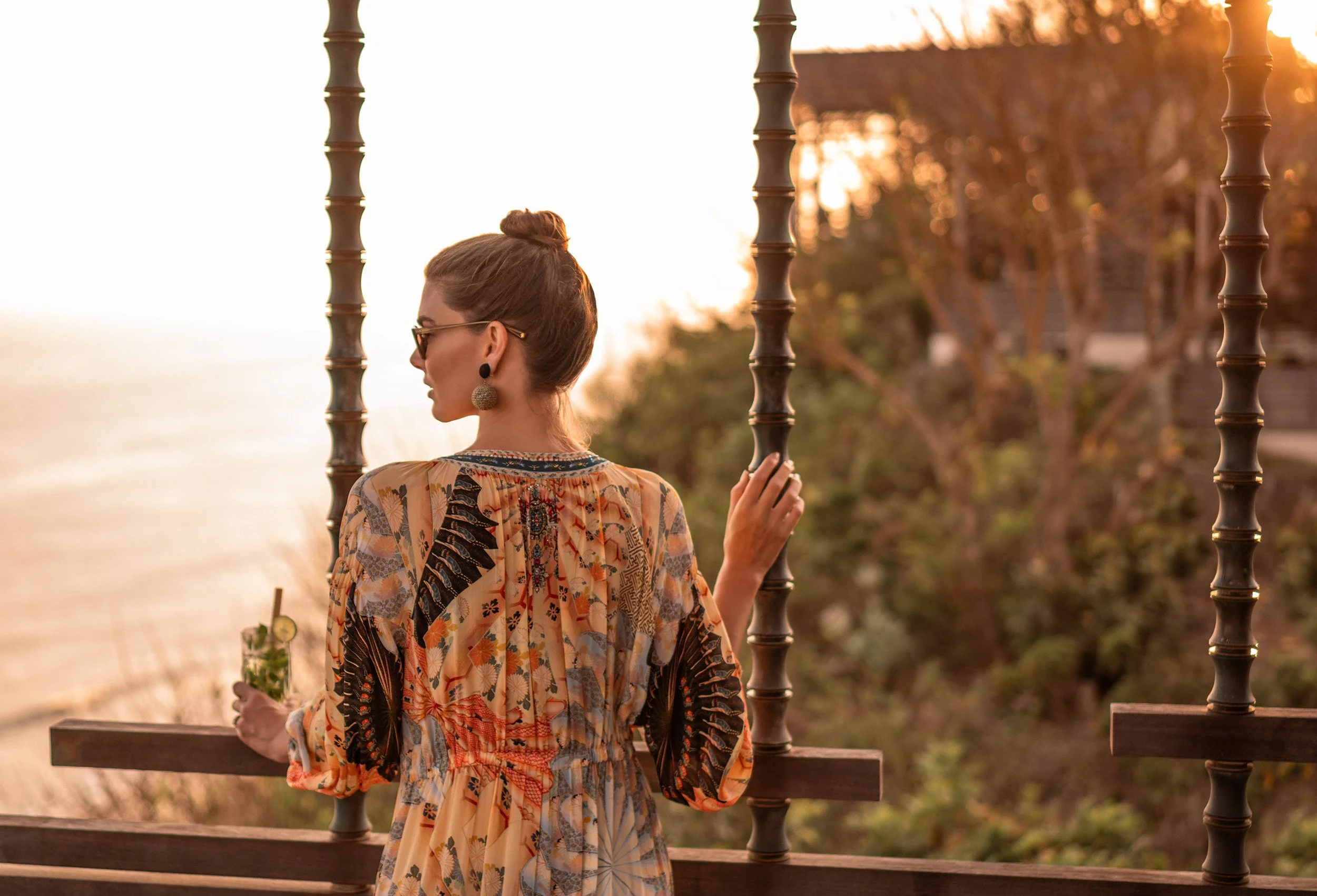 Woman with brown hair in a bun, wearing sunglasses, earrings, and a colorful patterned dress, sitting on a bench by a body of water during sunset.
