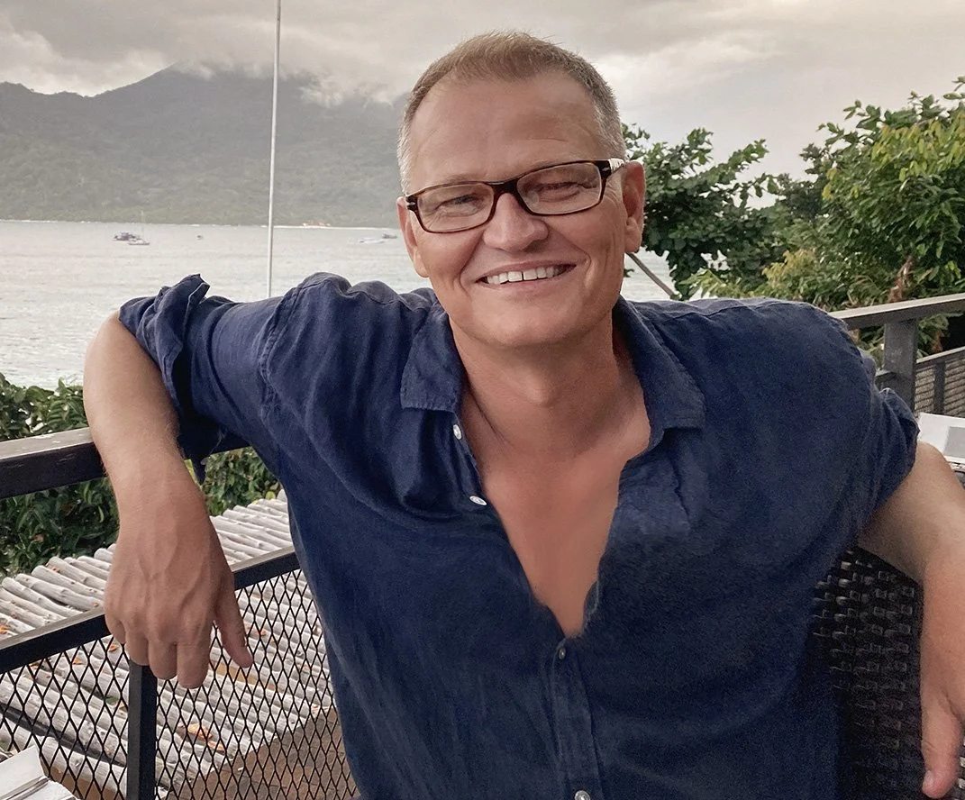 A smiling man with glasses and short hair sitting outdoors on a balcony, with a lake and mountains in the background.
