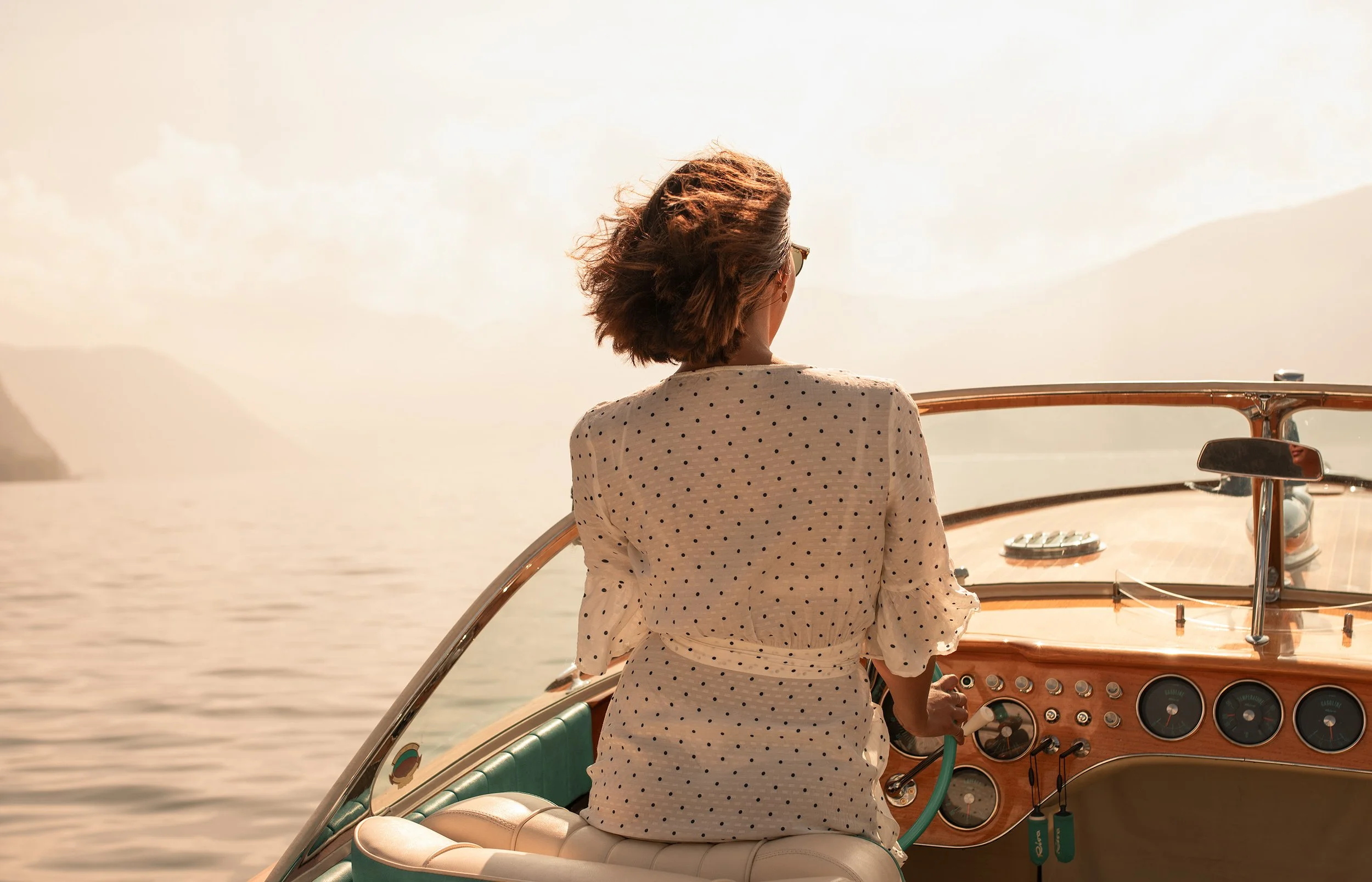A woman with short brown hair wearing sunglasses and a white dress with black polka dots sailing on a boat at sunset.