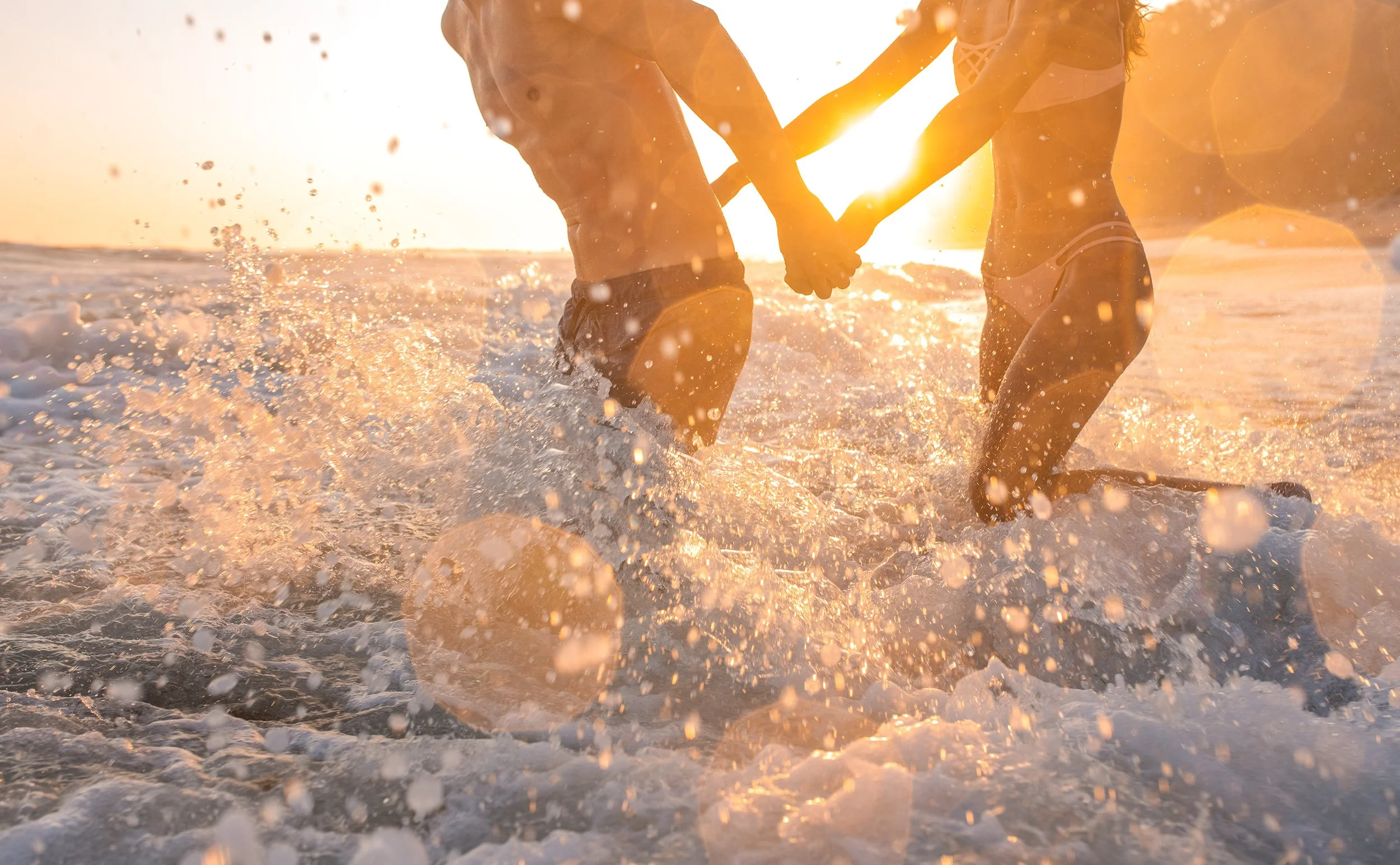 Couple holding hands while running through the ocean at sunset, splashing water, with the sun low on the horizon creating a warm glow.