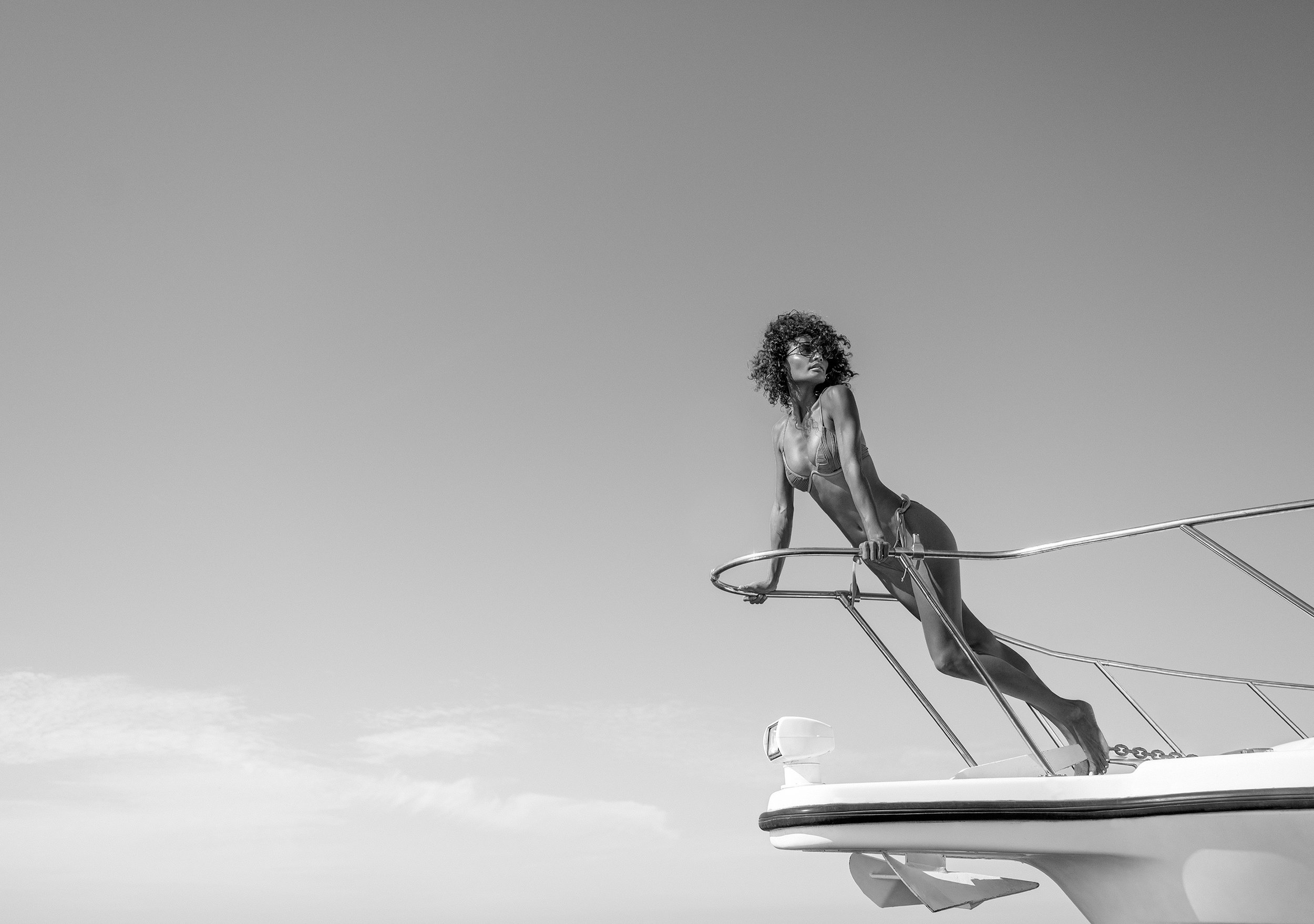 A woman with curly hair and sunglasses leaning on the railing of a boat or yacht, looking into the distance against a clear sky. Photographed by Kelly Fajack. Fashion, resort, luxury lifestyle photographer. 
