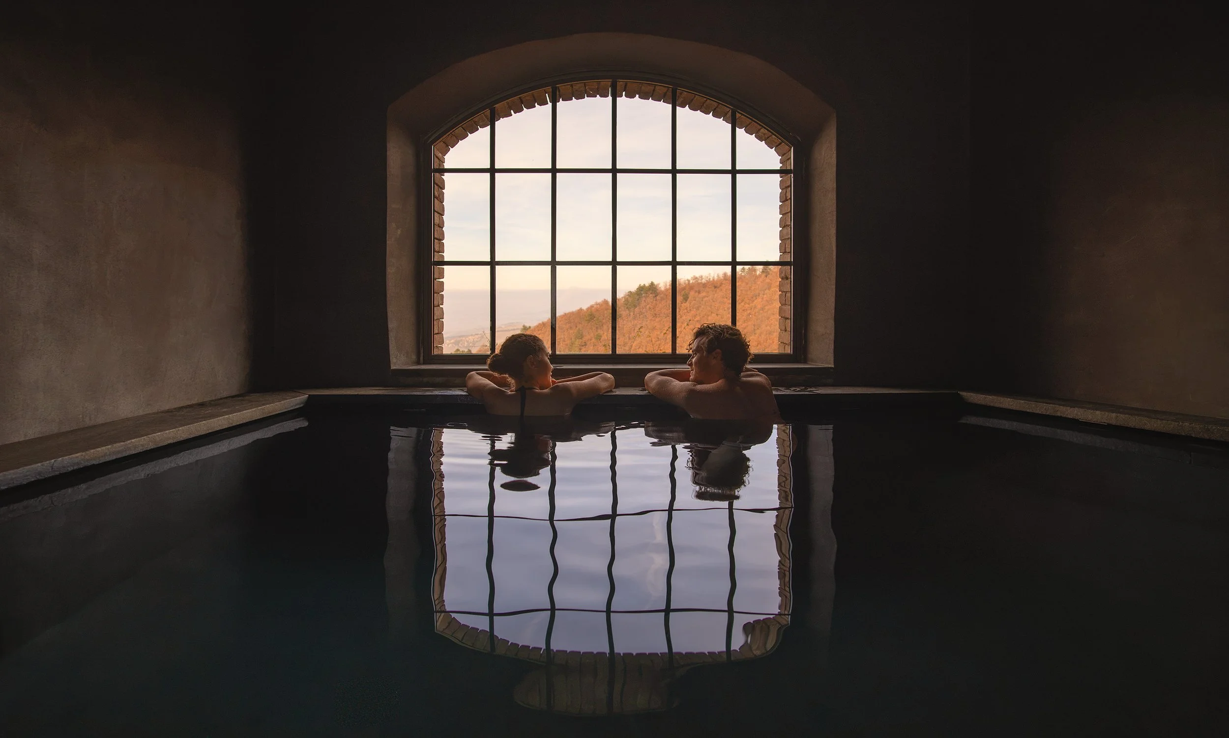 Two people are relaxing in a hot tub or bath inside a room with a large arched window showing a scenic view of hills and a partly cloudy sky.