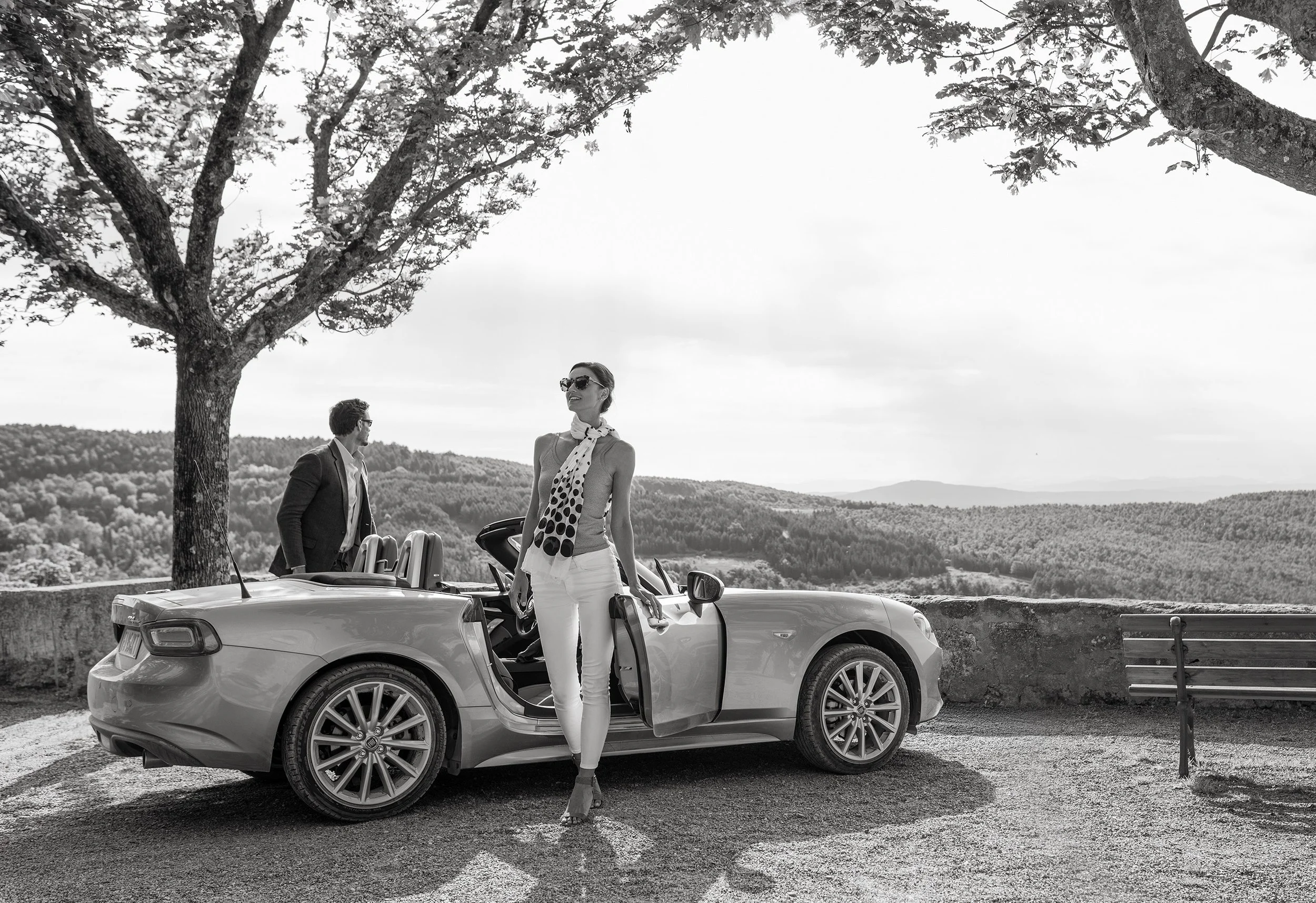 A woman in sunglasses and white pants stands beside a convertible car with two men in the background, all outdoors with rolling hills and trees.
